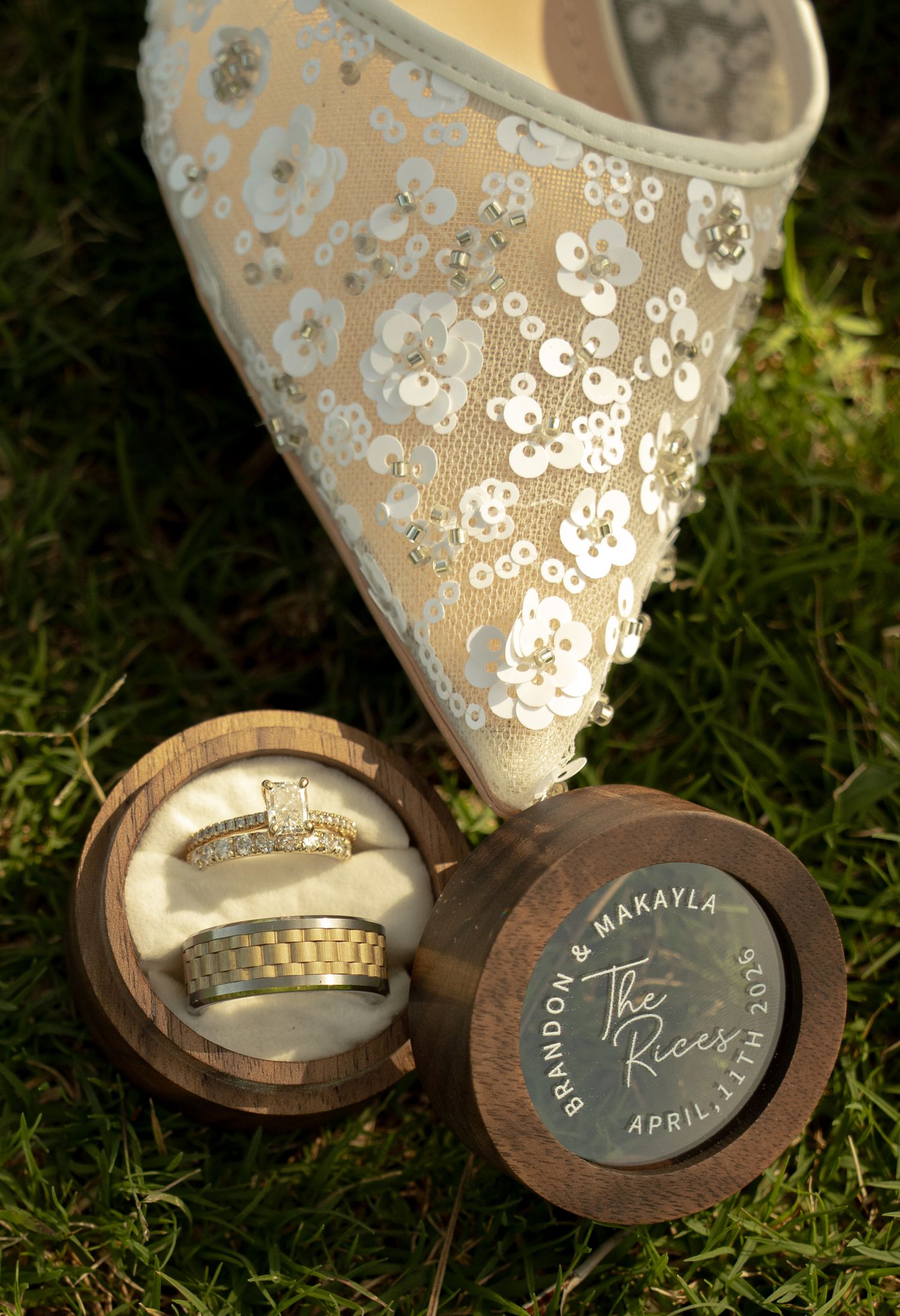 Personalized wooden ring box engraved with Brandon & Makayla The Rices April 17th beside a sequined wedding shoe on grass.