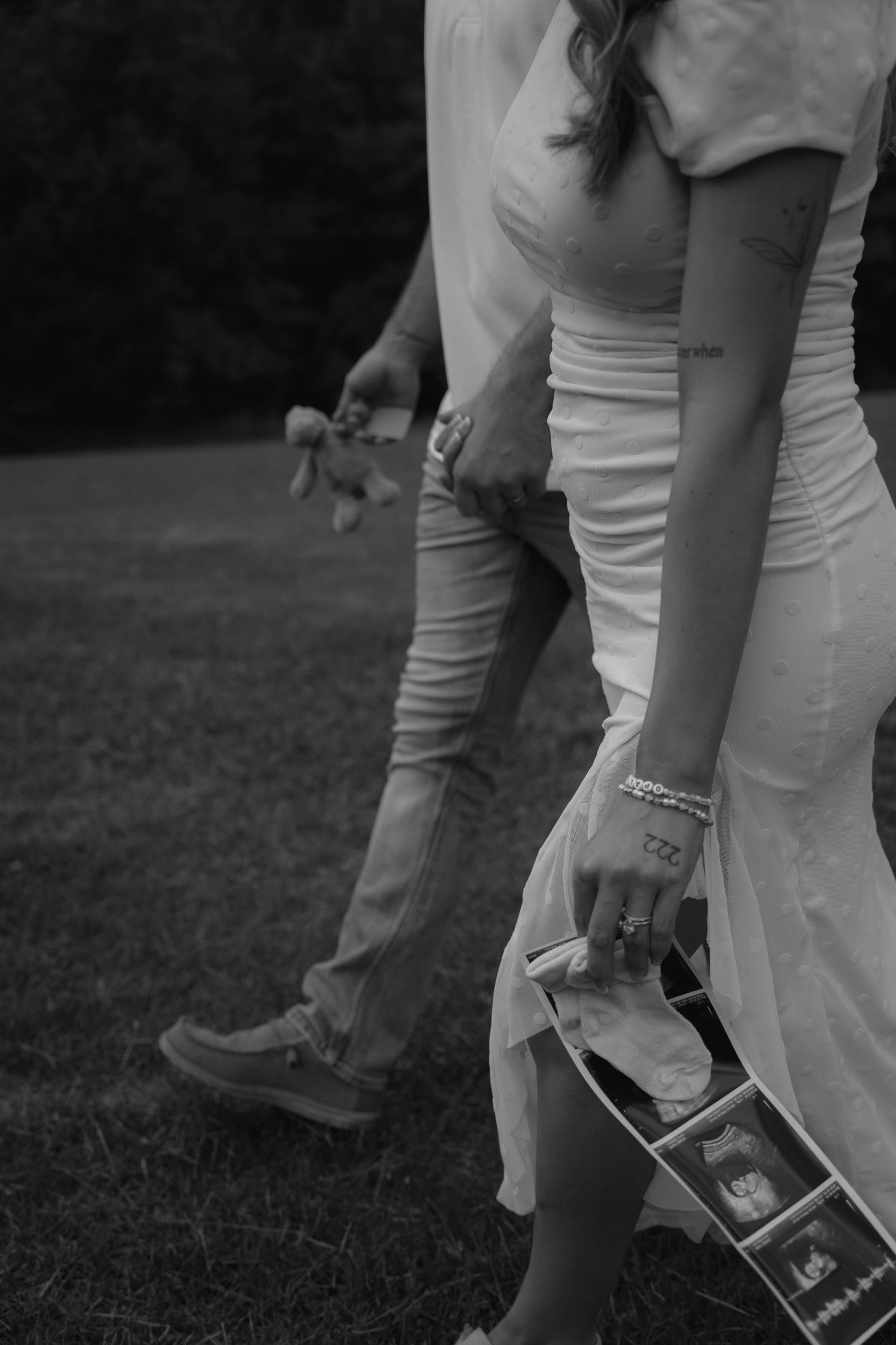 Black and white photo of a couple walking outdoors, woman holding ultrasound photos and baby shoes, pregnancy announcement.