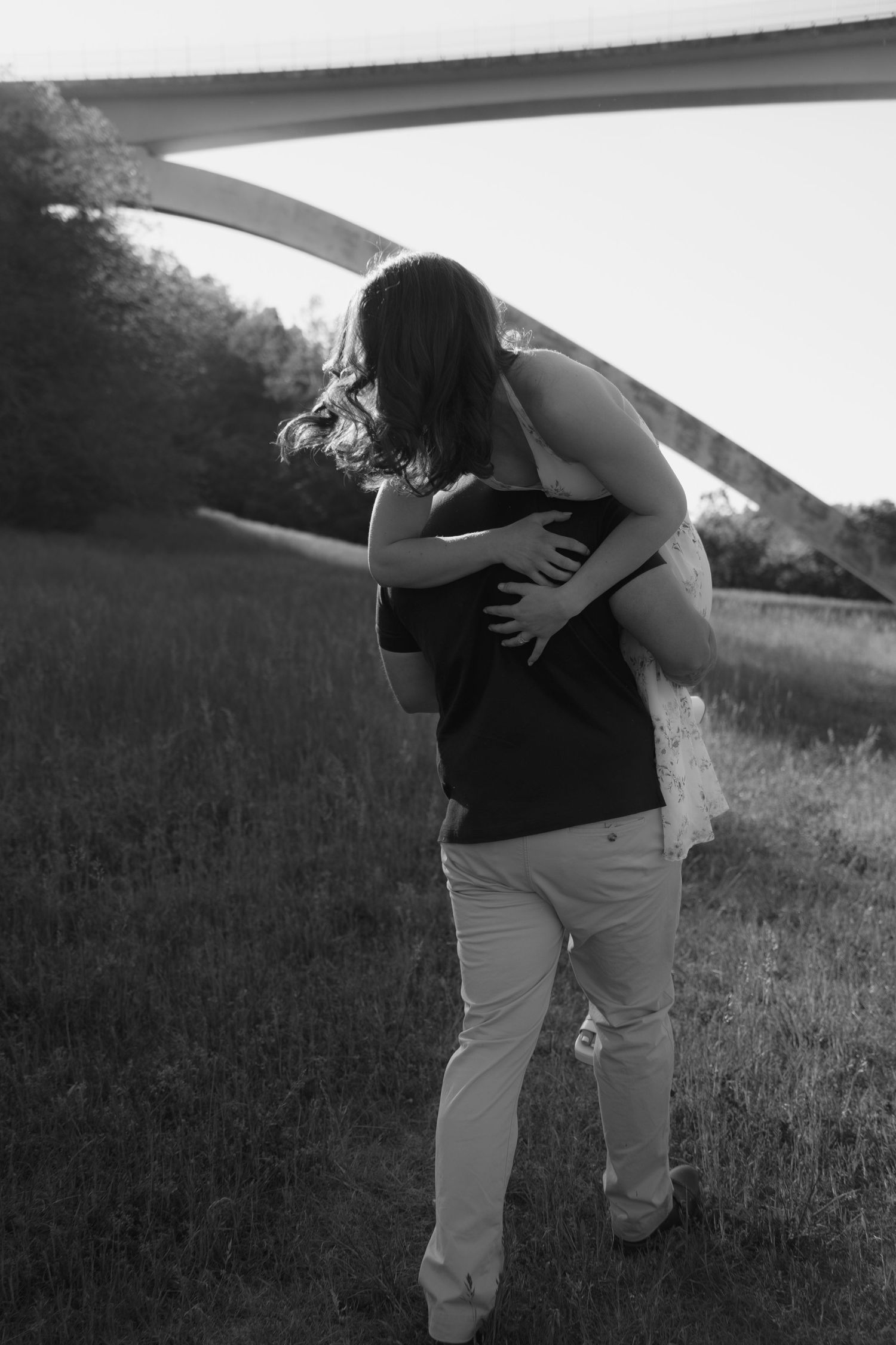Black and white photo of a couple embracing in a field near an arched bridge surrounded by trees.
