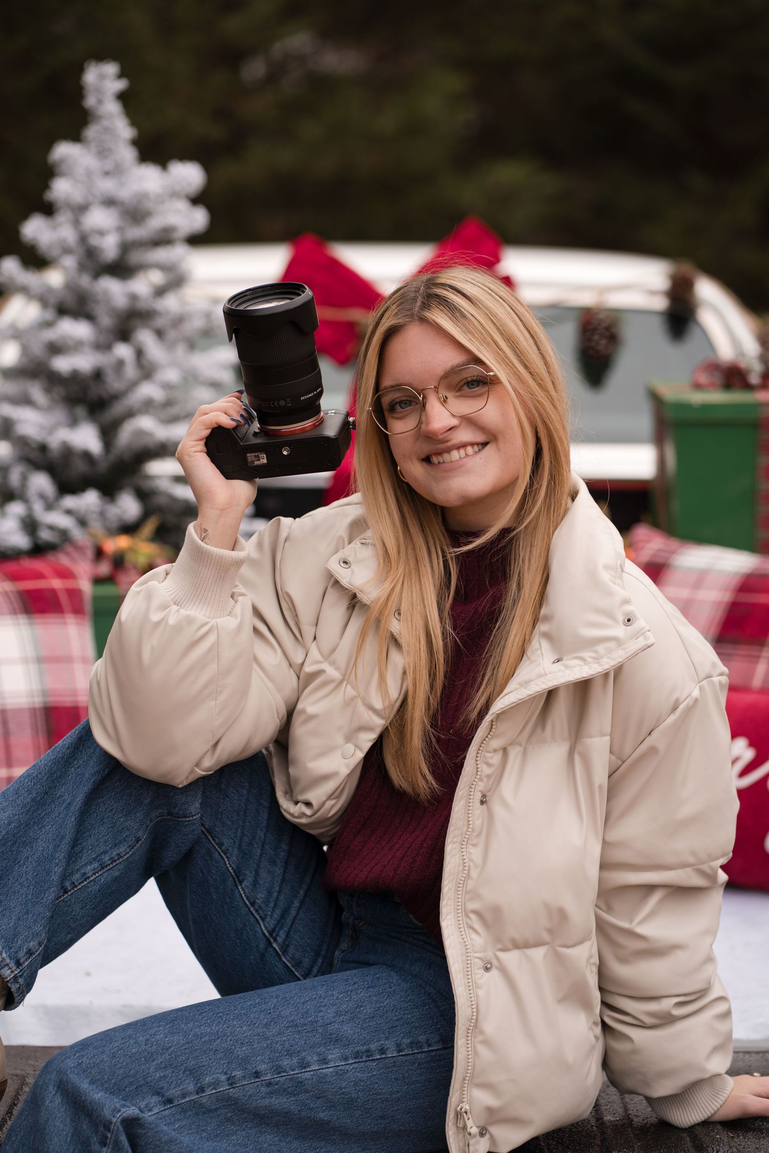 A woman in a cream winter coat and glasses poses with a camera in a festive holiday setting with snow-covered trees.