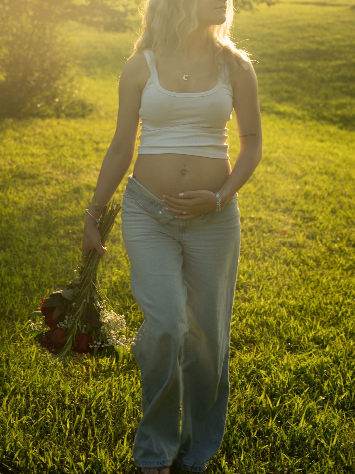 Pregnant woman in white crop top and light jeans holding red roses in sunlit green field.