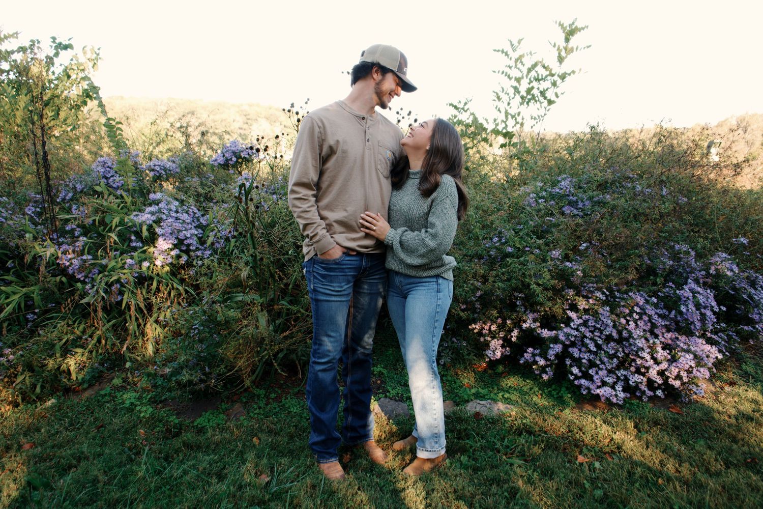 Couple standing together in a field during golden hour with wildflowers and natural scenery in the background.