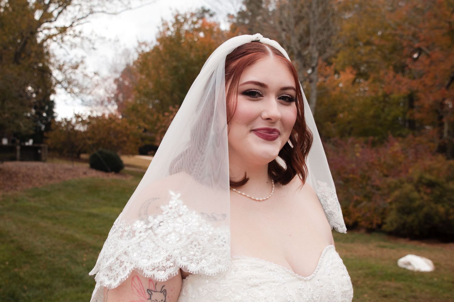 A bride in a white lace wedding dress and veil poses outdoors during autumn with colorful fall foliage in the background.
