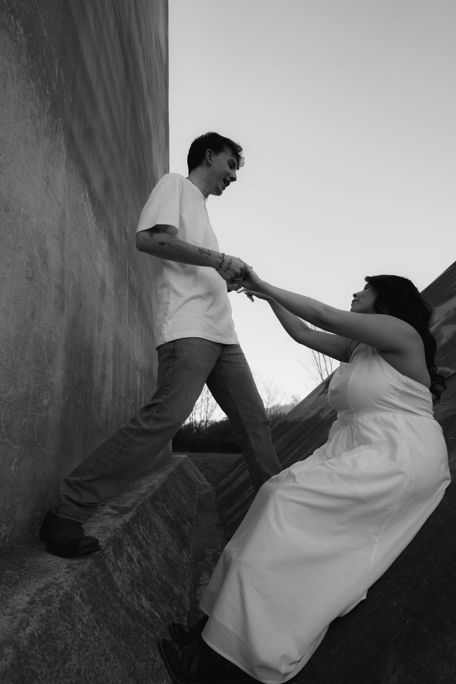 Black and white photo of a couple holding hands, man helping woman in white dress up stone steps outdoors.
