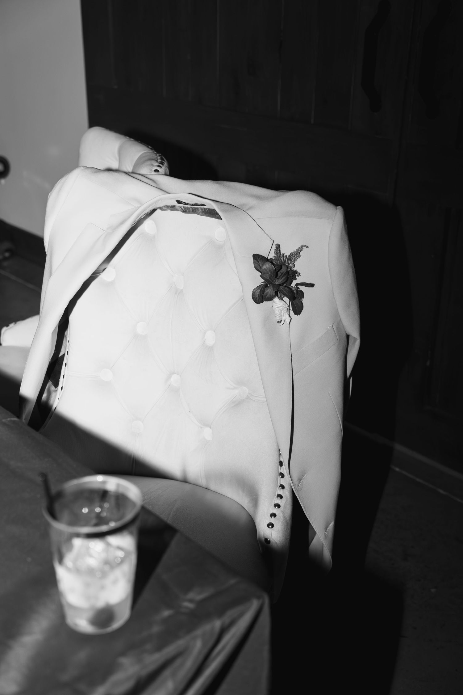 Black and white photo of a white tuxedo jacket with boutonniere draped over a tufted chair at a wedding reception.