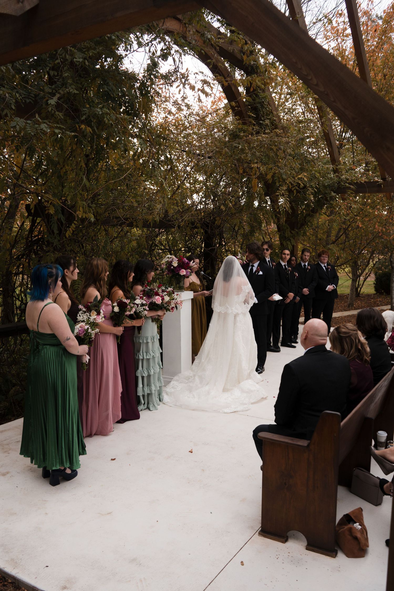 Wedding ceremony setup outdoors with wooden pews and guests seated under autumn trees.