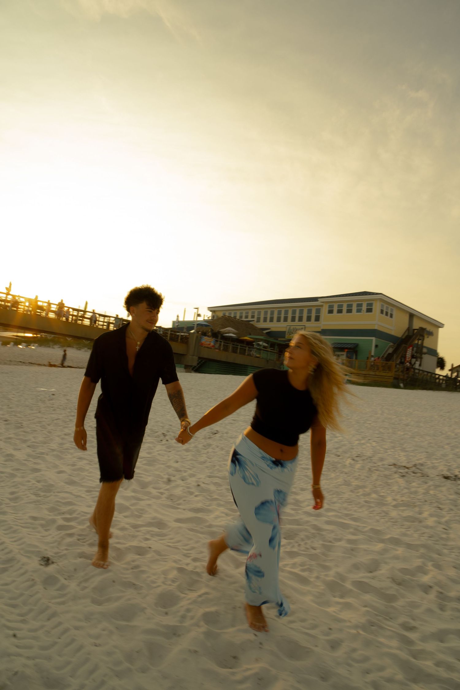 A young couple holds hands and runs barefoot on a white sandy beach at sunset near a coastal building.