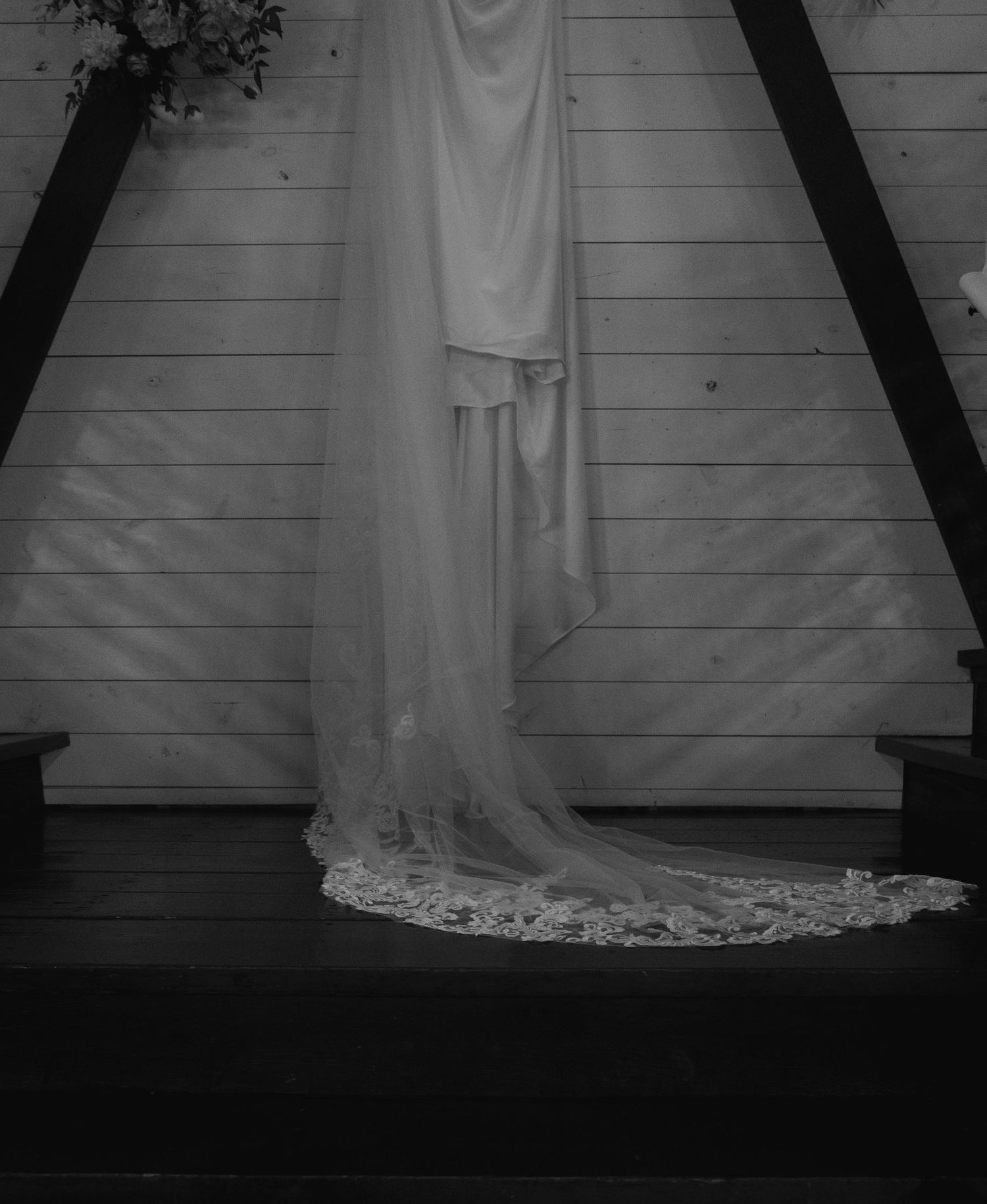 Black and white photo of an elegant wedding dress with lace-trimmed cathedral veil displayed in a rustic wooden chapel.