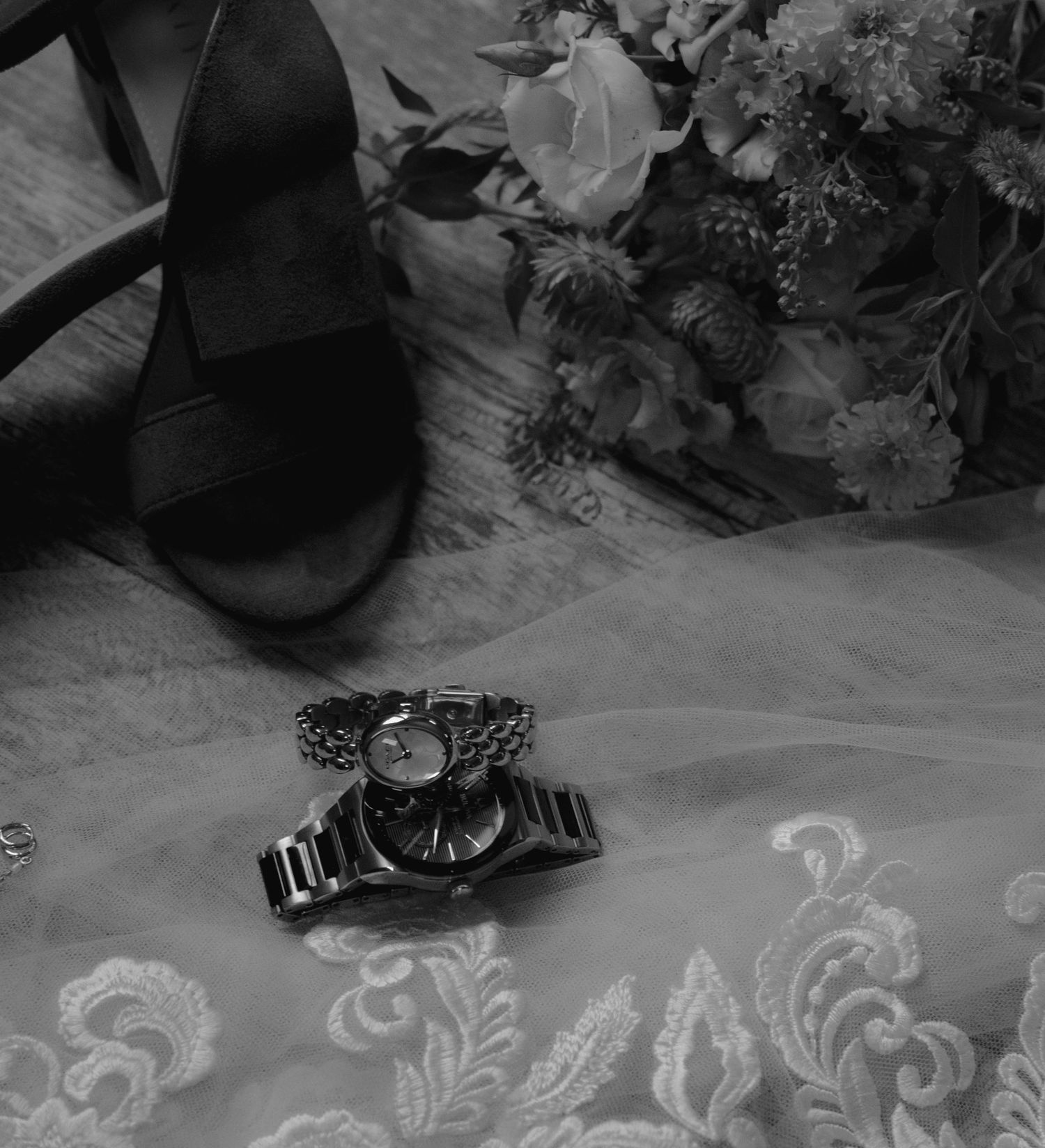 Black and white flat lay of a wristwatch, heeled sandal, floral bouquet, and lace fabric for a wedding.