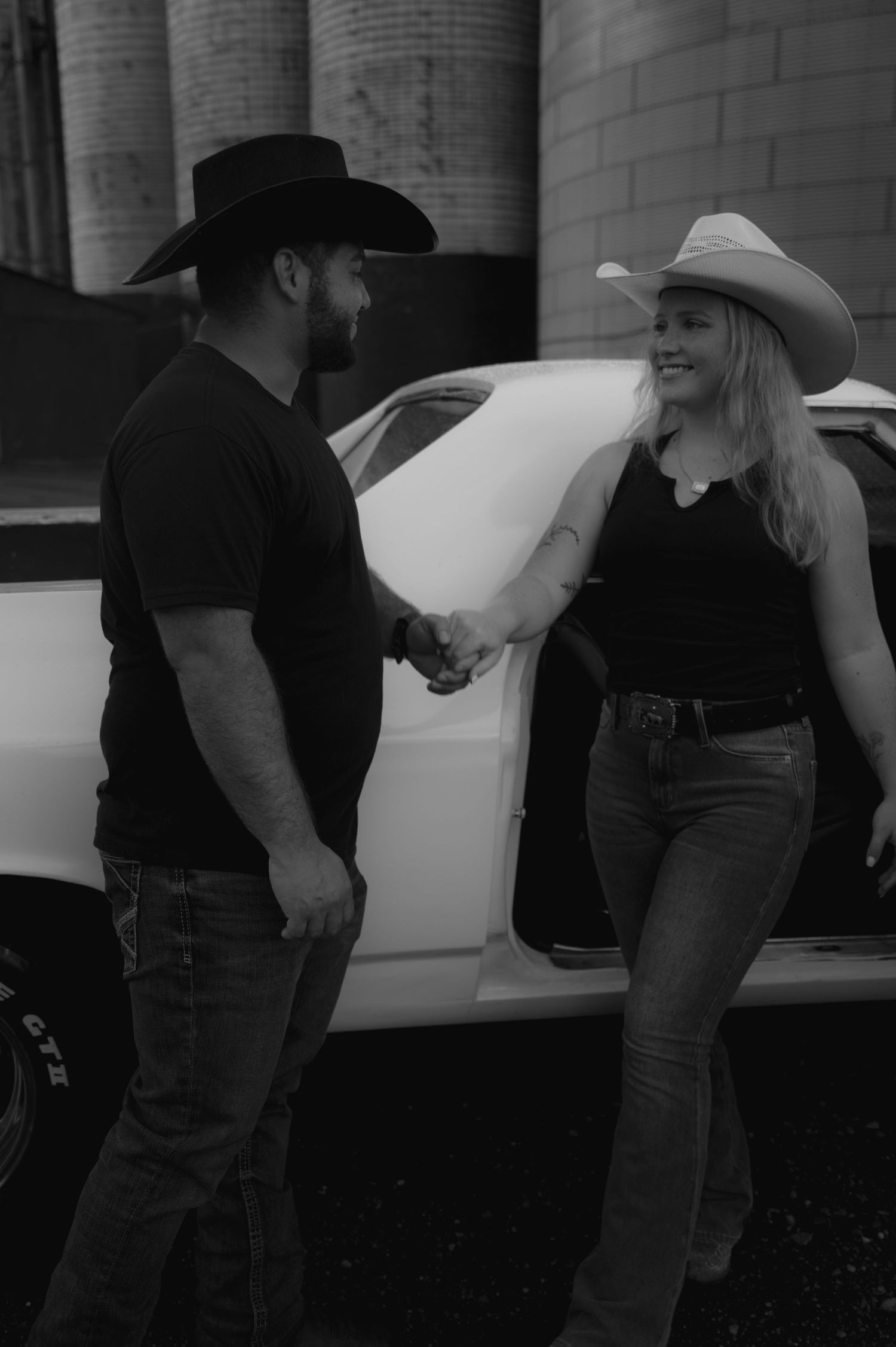 Black and white photo of a couple in cowboy hats holding hands beside a classic white car.