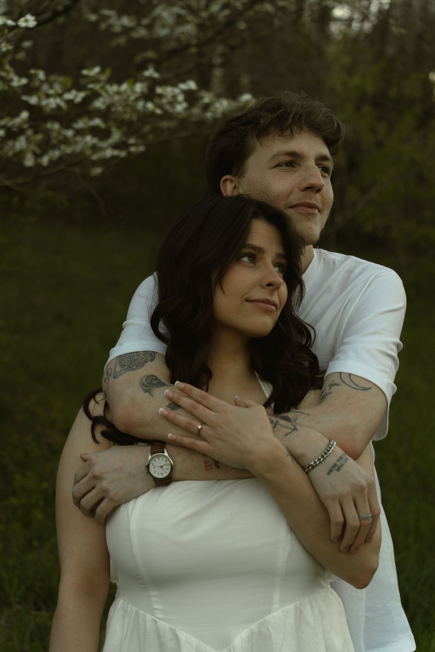 A couple embraces in a lush garden, man in white shirt wraps arms around tattooed woman in white dress near blooming trees.