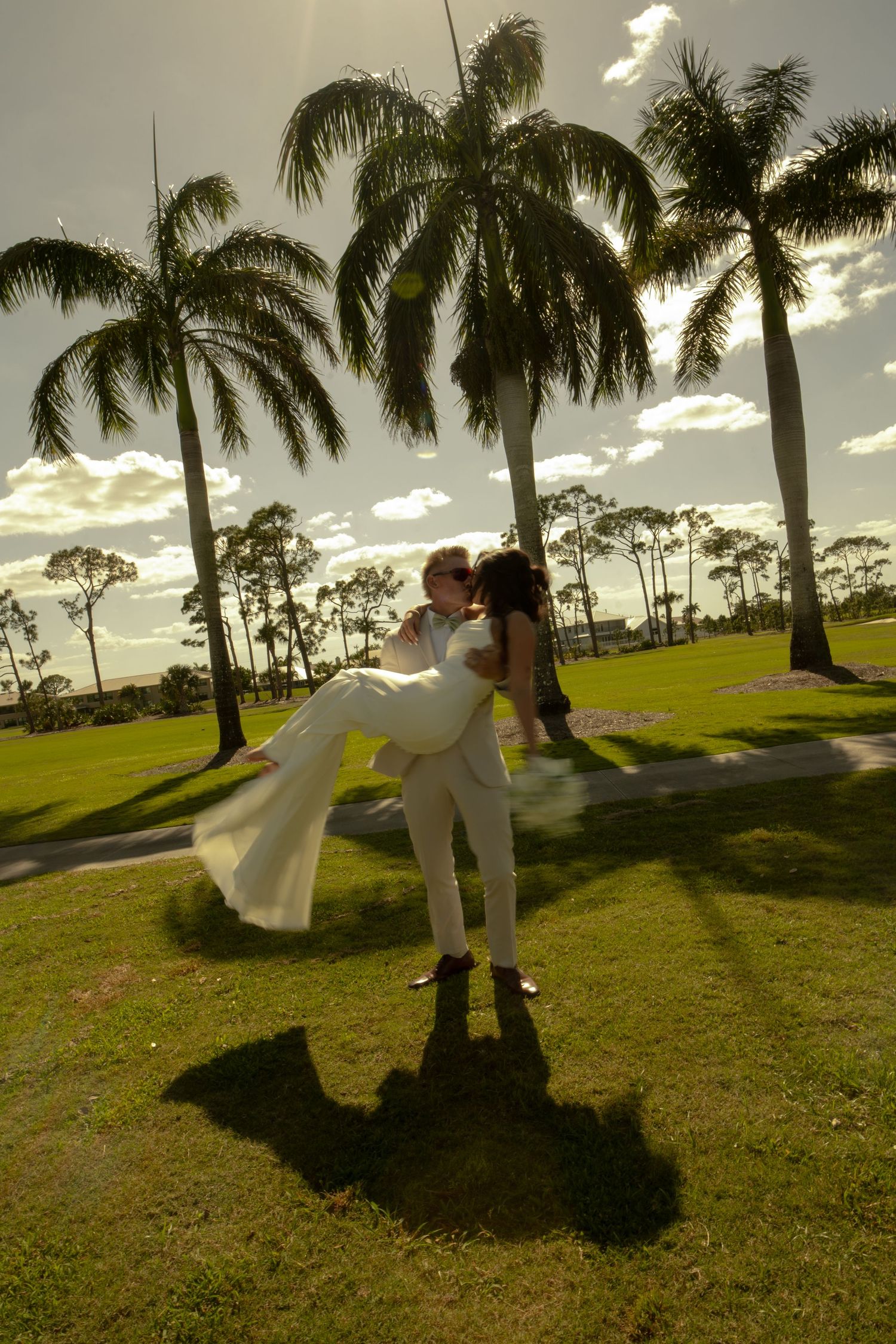 Groom lifting bride in white dress among tropical palm trees on sunny green lawn at outdoor wedding celebration.