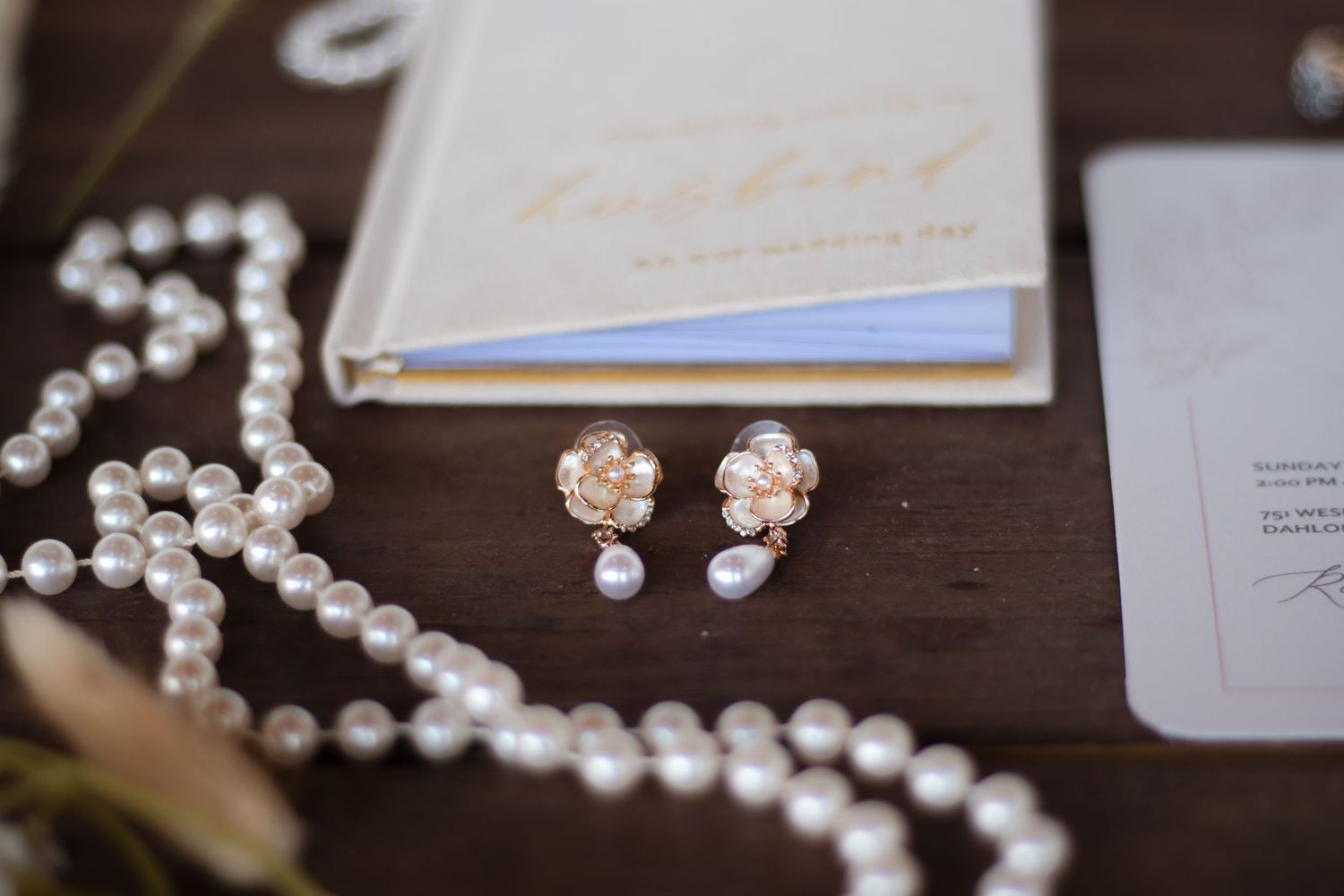 Wedding details featuring delicate pearl necklaces and invitation cards arranged on a dark wooden surface.
