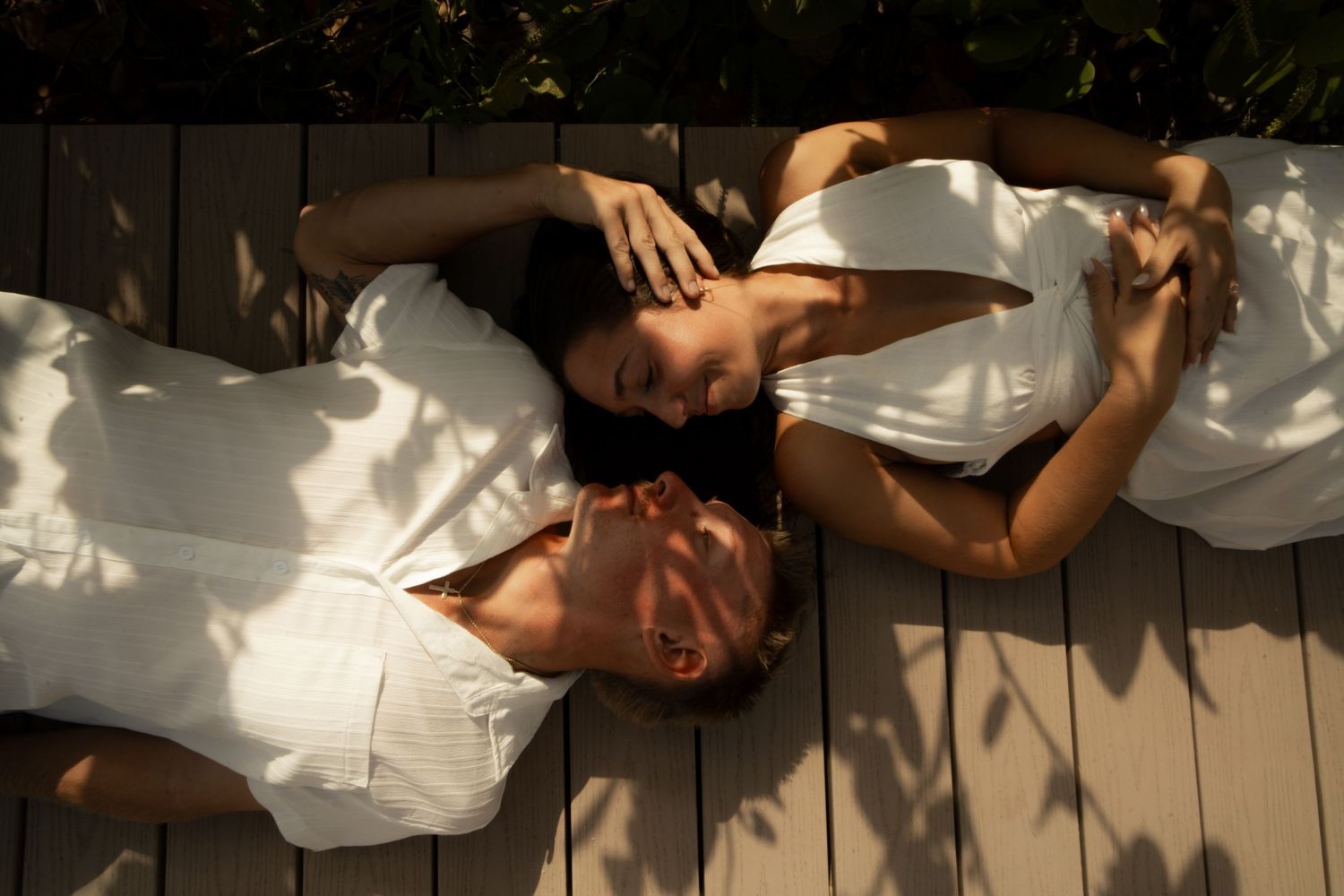 Two women in white dresses lying on wooden decking in dappled sunlight, relaxing outdoors together.