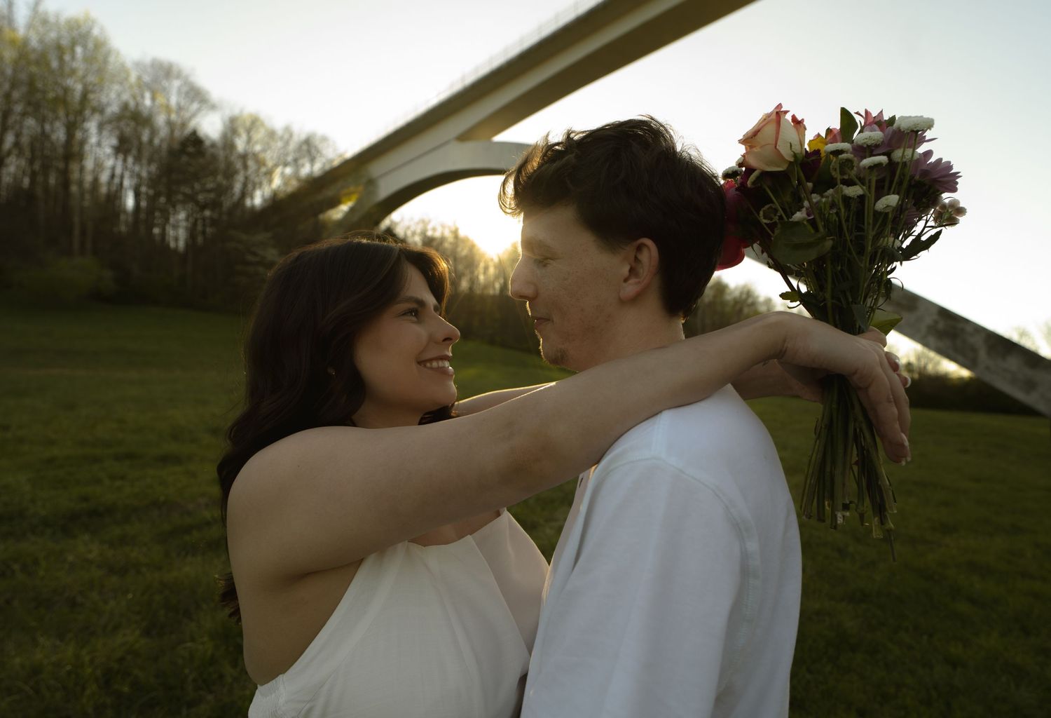 A couple shares a romantic moment outdoors, woman in white dress holds flowers while smiling at her partner during golden hour.