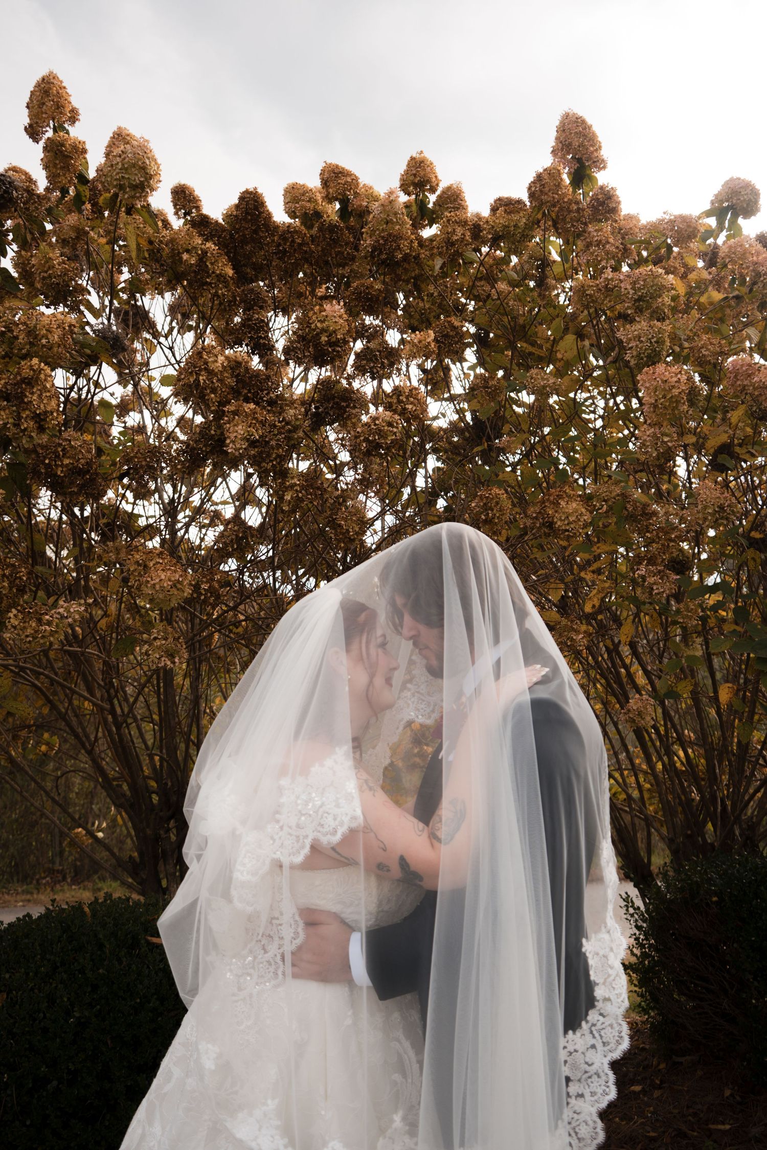 A romantic wedding moment captured under autumn trees with the couple embracing beneath a flowing veil.