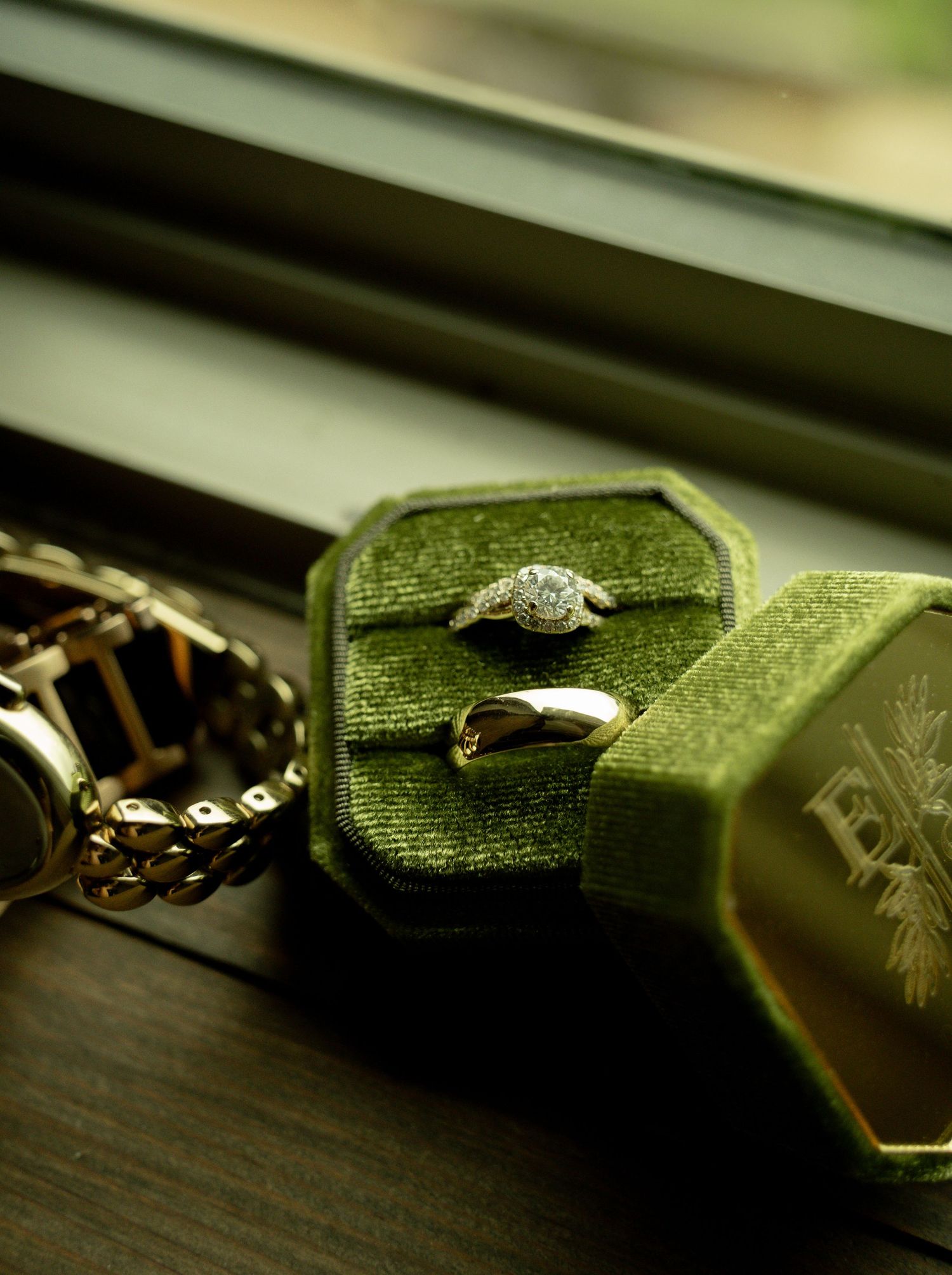 Diamond engagement ring and gold wedding band displayed in a green velvet hexagonal ring box on a windowsill.