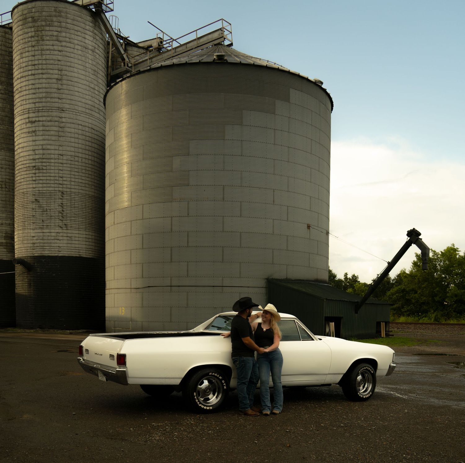Couple embracing beside a white classic El Camino in front of large grain silos on a farm.