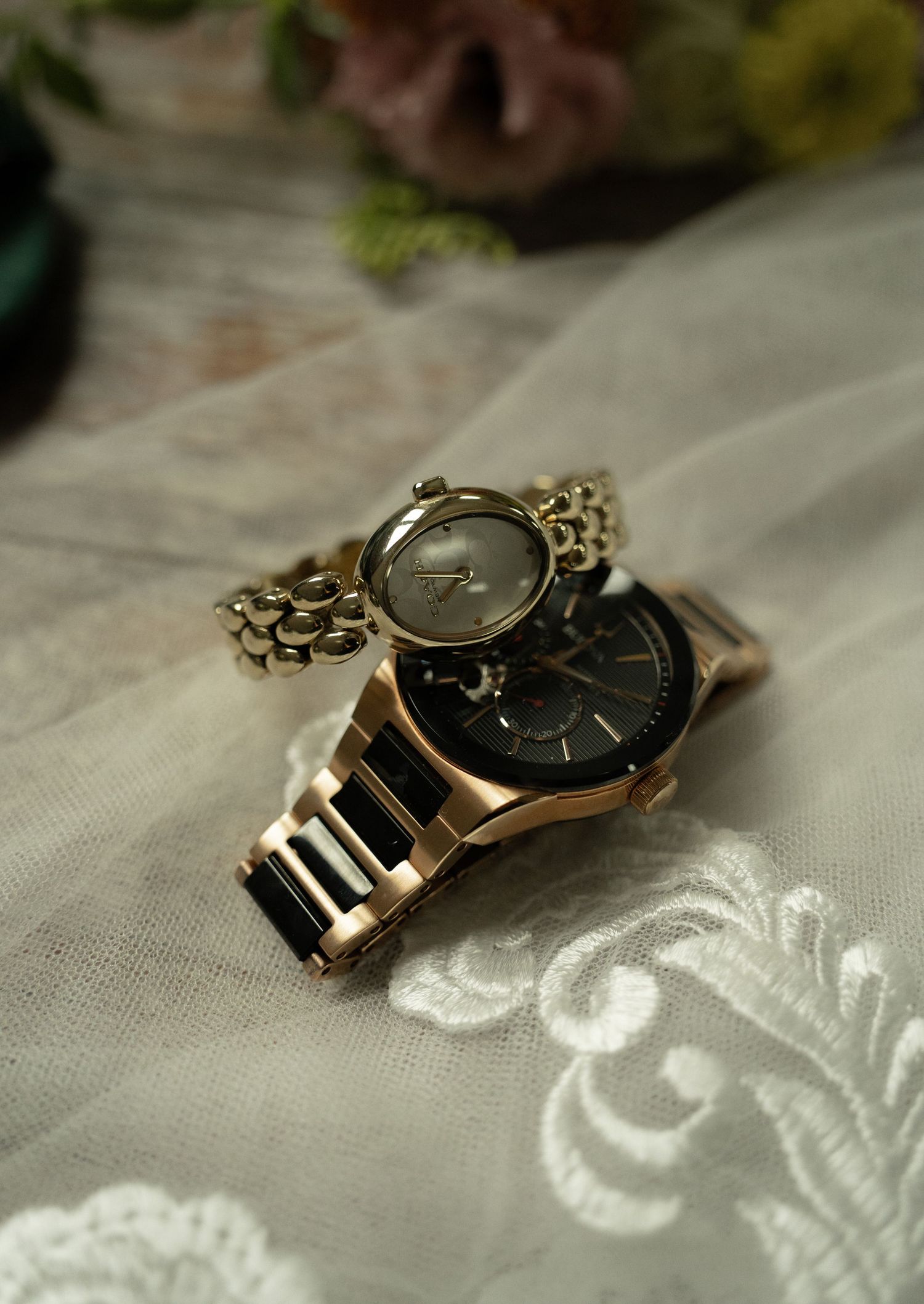 Two gold and black luxury wristwatches stacked on white lace fabric with floral decor in background.