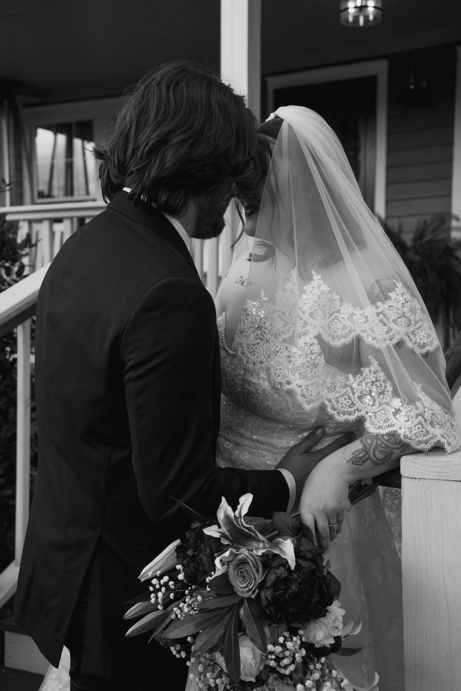 A wedding couple embraces near white columns, with the bride wearing a lace-trimmed veil.