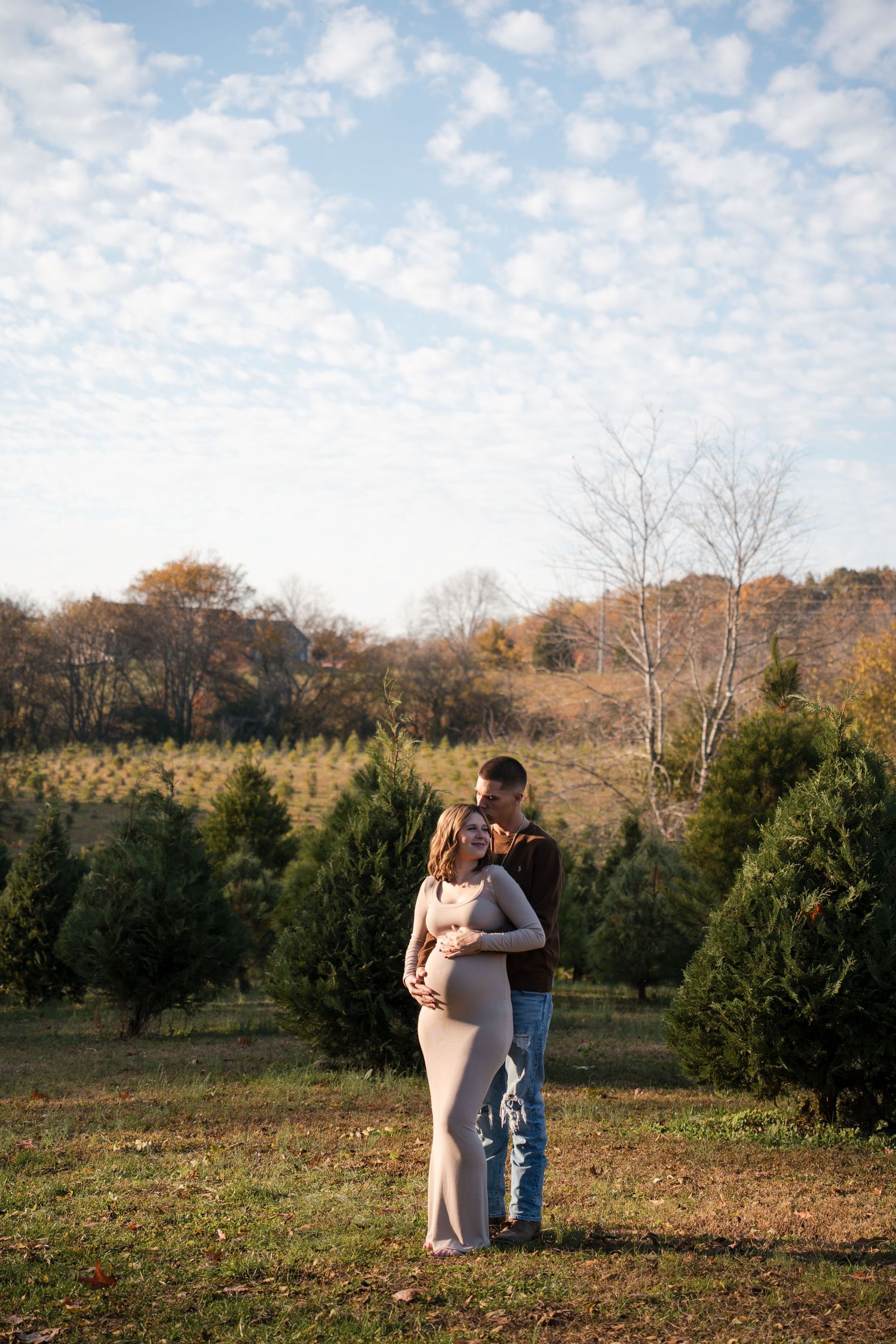 A couple embraces in a scenic Christmas tree farm on an autumn afternoon with golden light filtering through the trees.