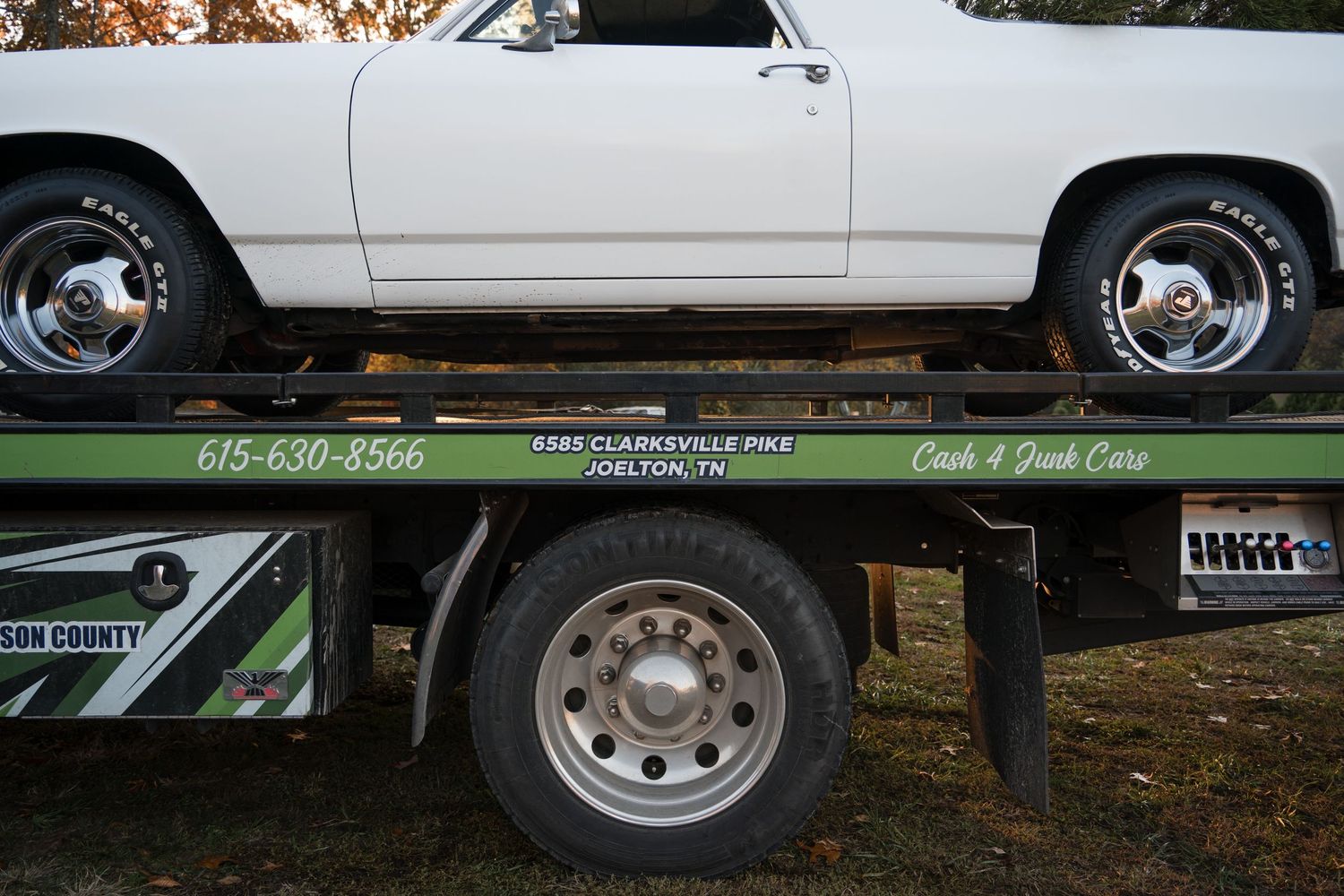 White classic car being transported on a green flatbed tow truck with visible wheel details.