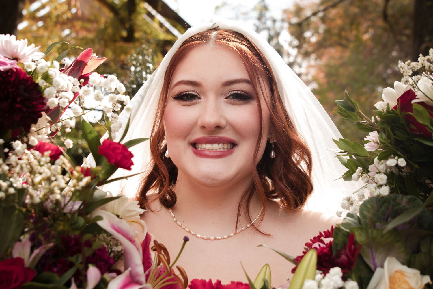 A bride smiles radiantly while surrounded by deep red and white floral arrangements on her wedding day.