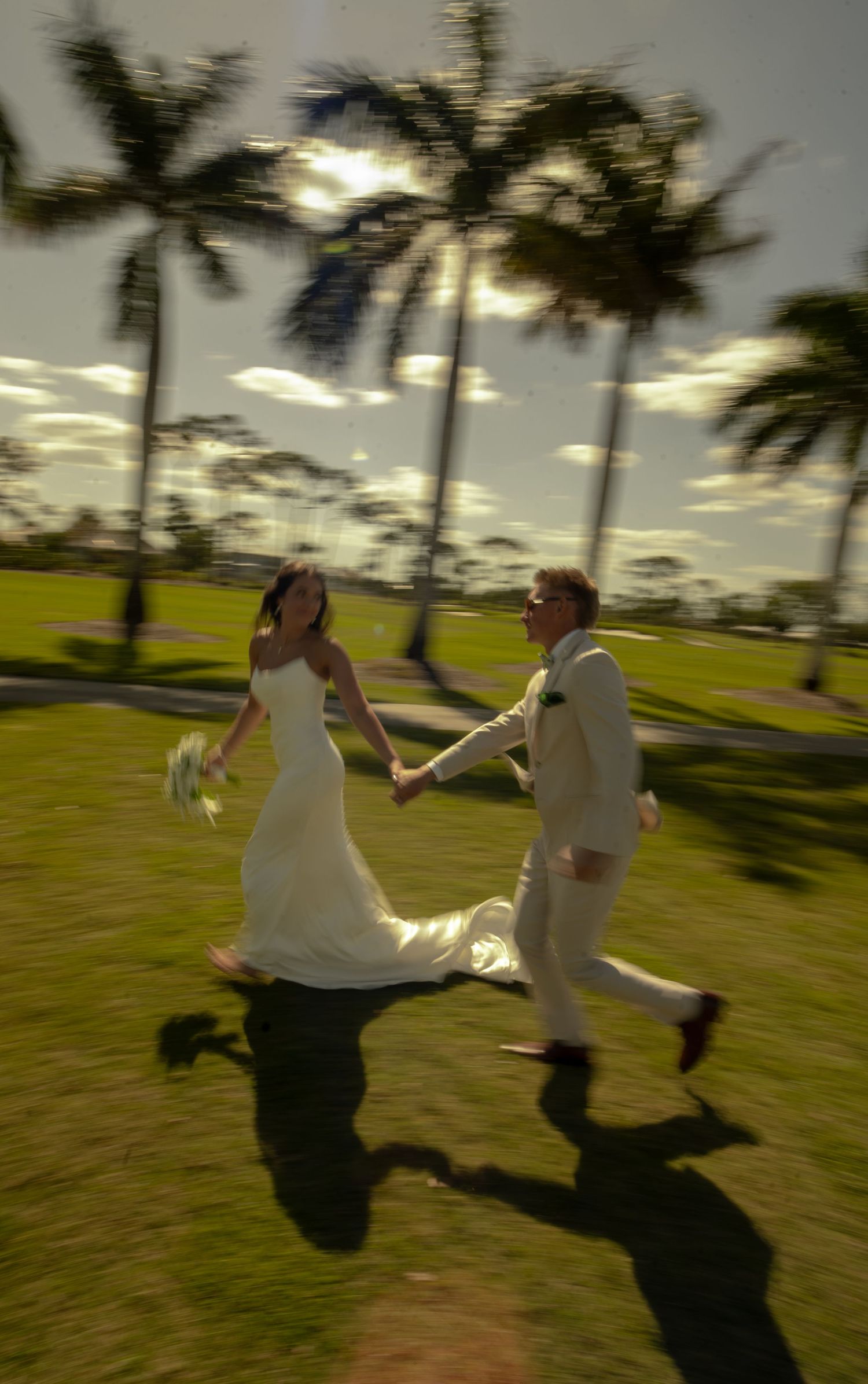 A joyful bride and groom run hand-in-hand across a lush green lawn surrounded by tall palm trees on their wedding day.
