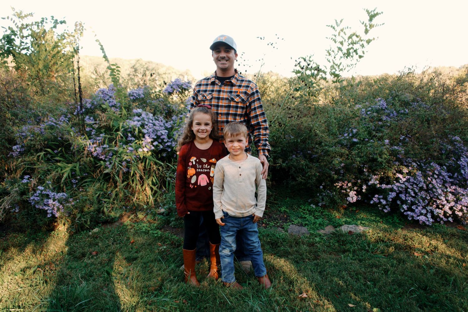 Three people stand together outdoors in front of blooming wildflowers on a sunny autumn day.