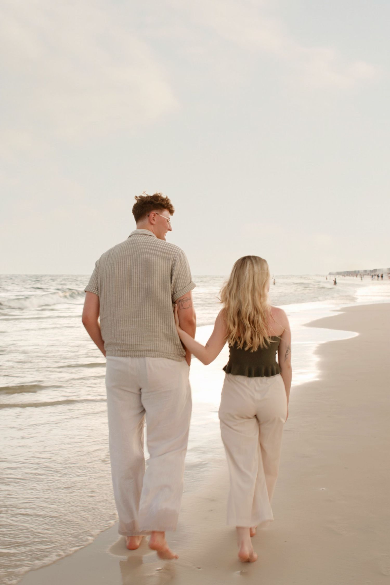 A couple wearing light colored clothing walks together barefoot along a serene beach at sunset.