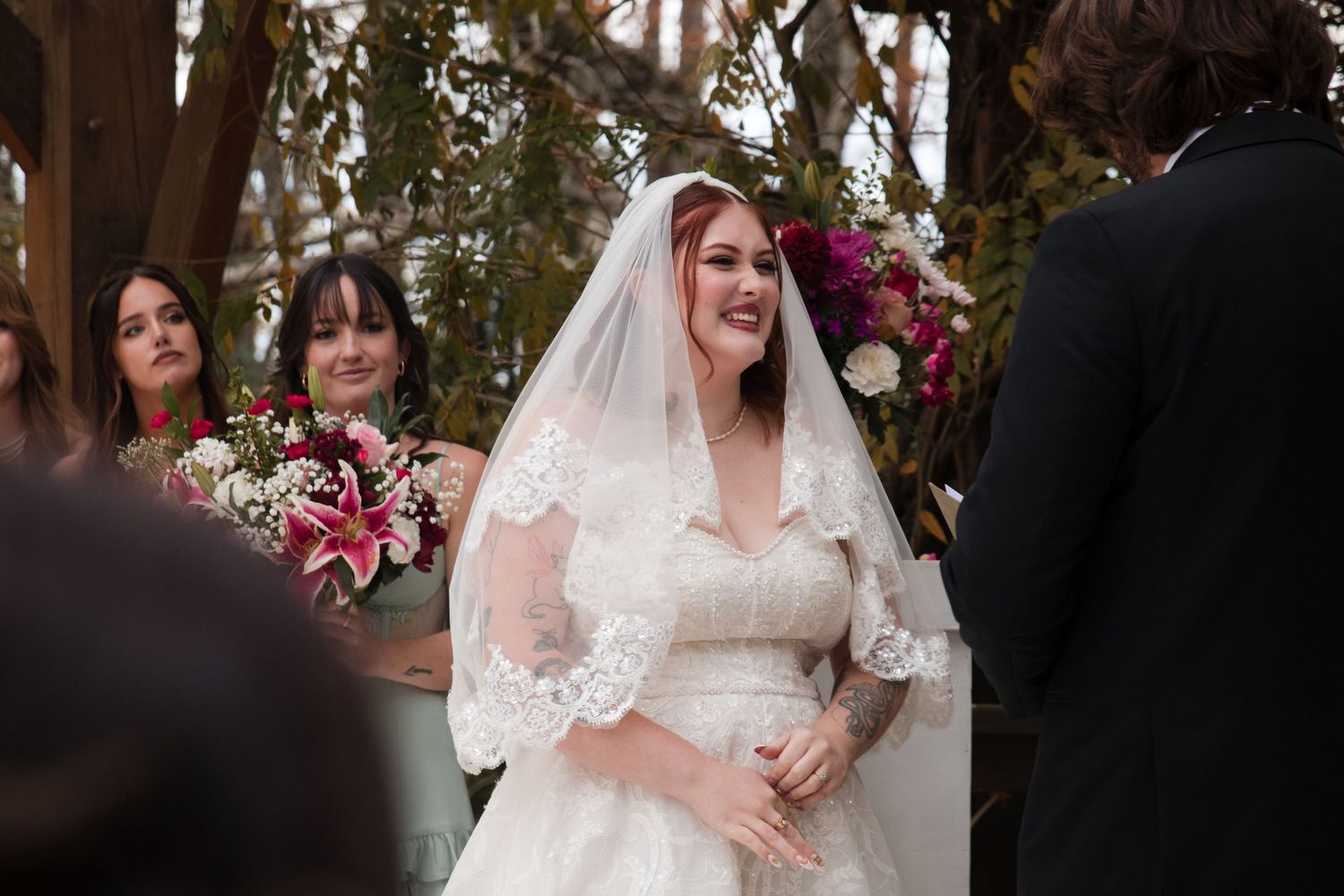 A bride in white lace dress poses with bridesmaids in a floral outdoor setting.
