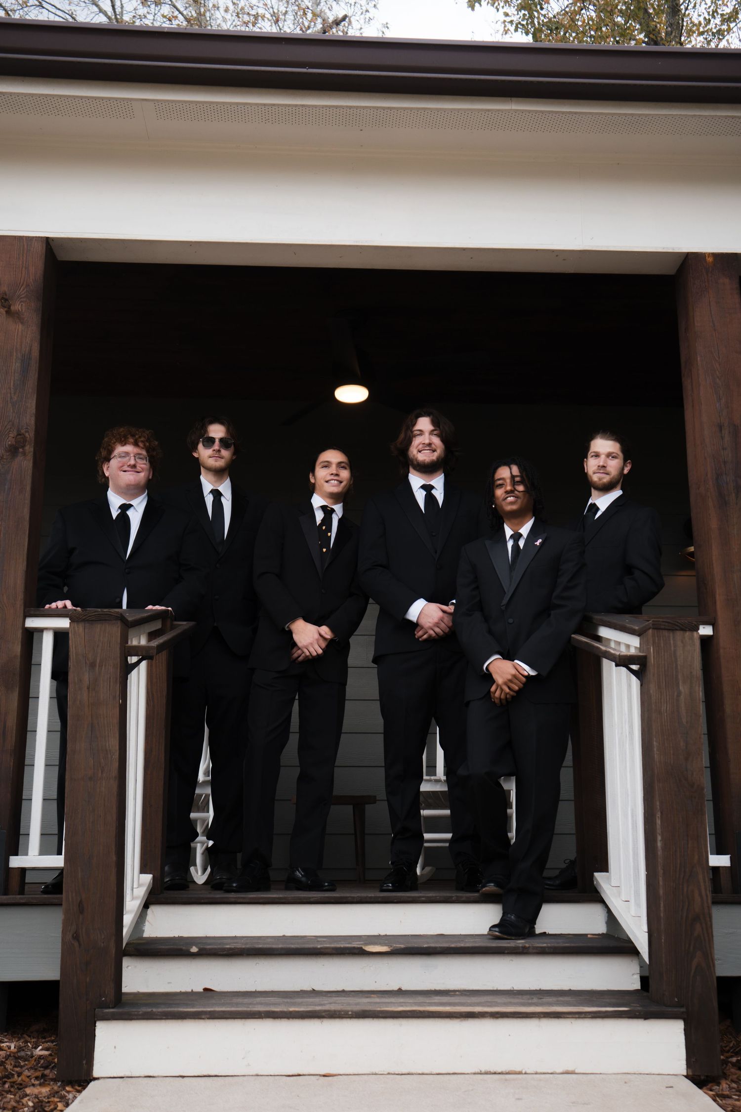 Groomsmen in black suits pose together on a white porch during a wedding photoshoot.