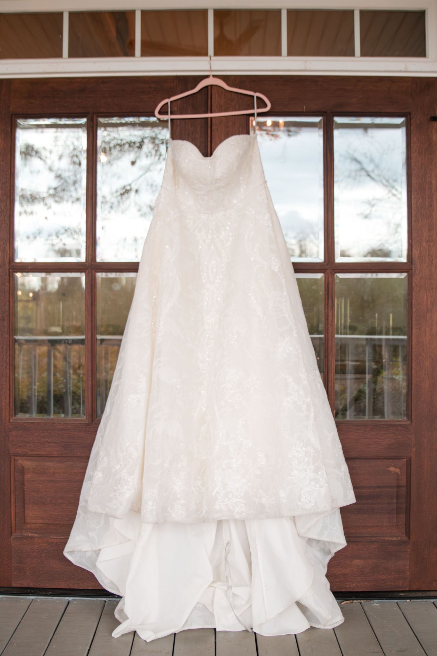 A white lace wedding dress hangs on a wooden hanger against rustic French doors with glass panes.