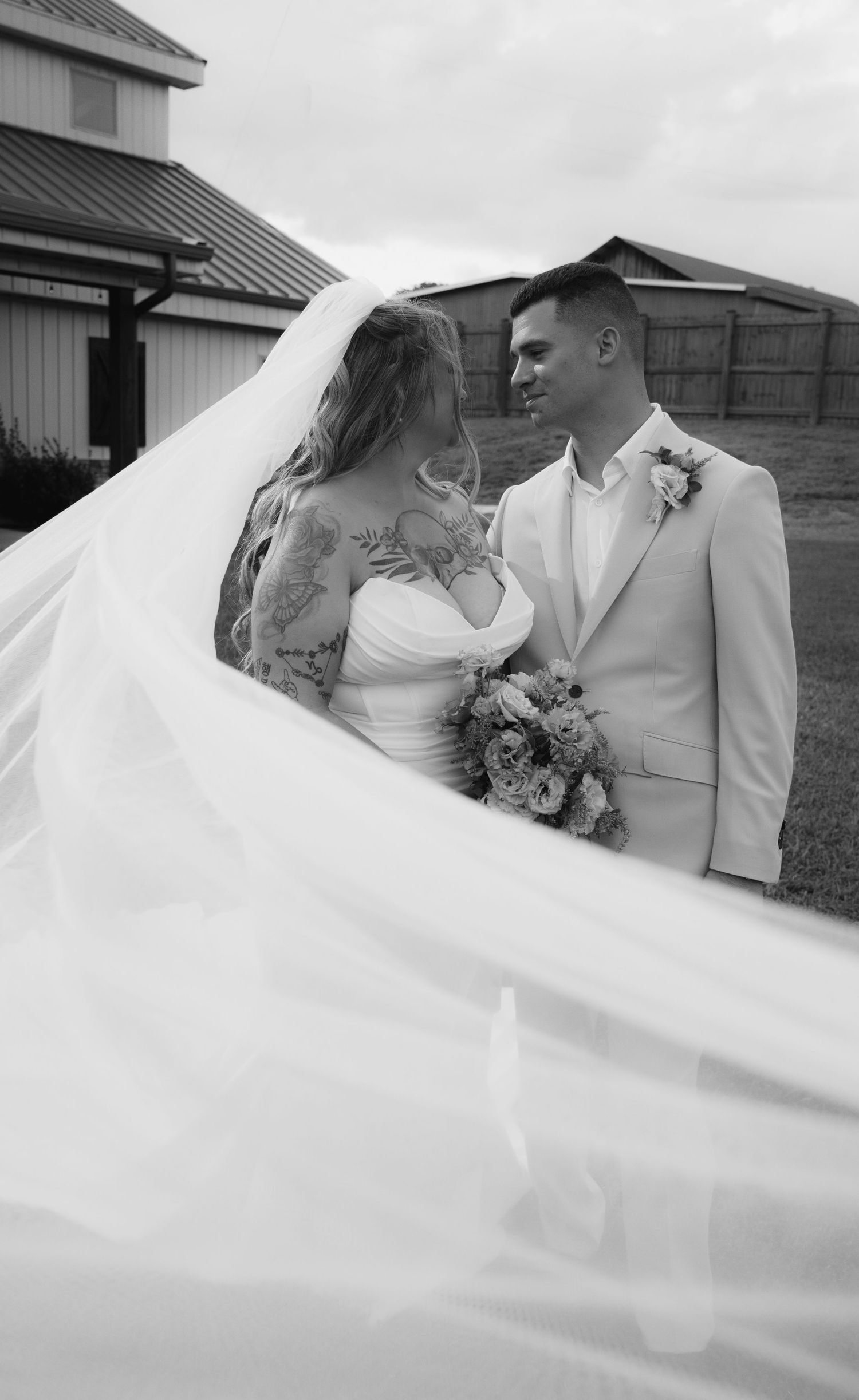 Bride and groom share a romantic moment as her veil flows in the wind near a barn.