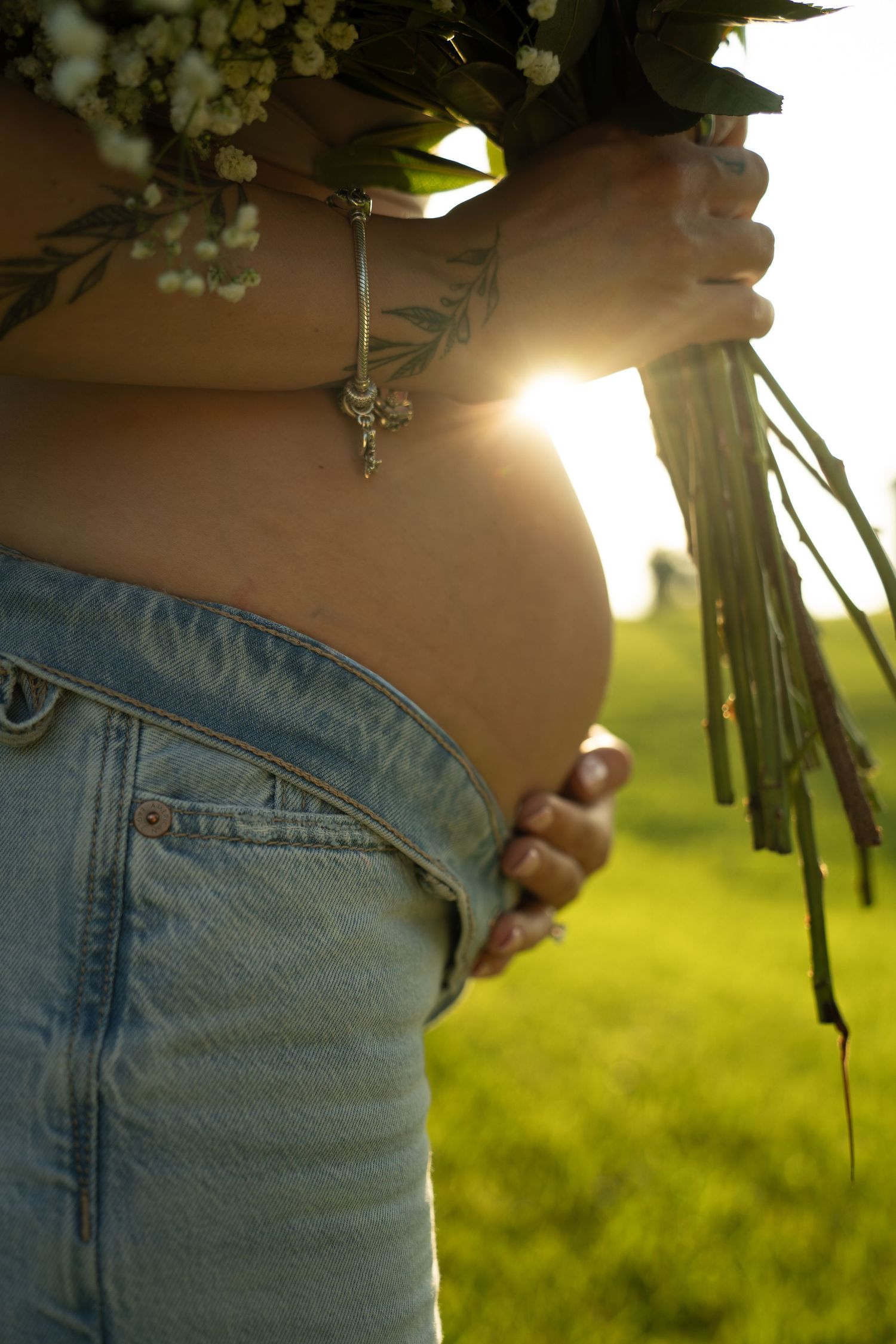 Pregnant woman holding wildflowers outdoors in a sunny green field, wearing jeans with exposed baby bump and a bracelet.