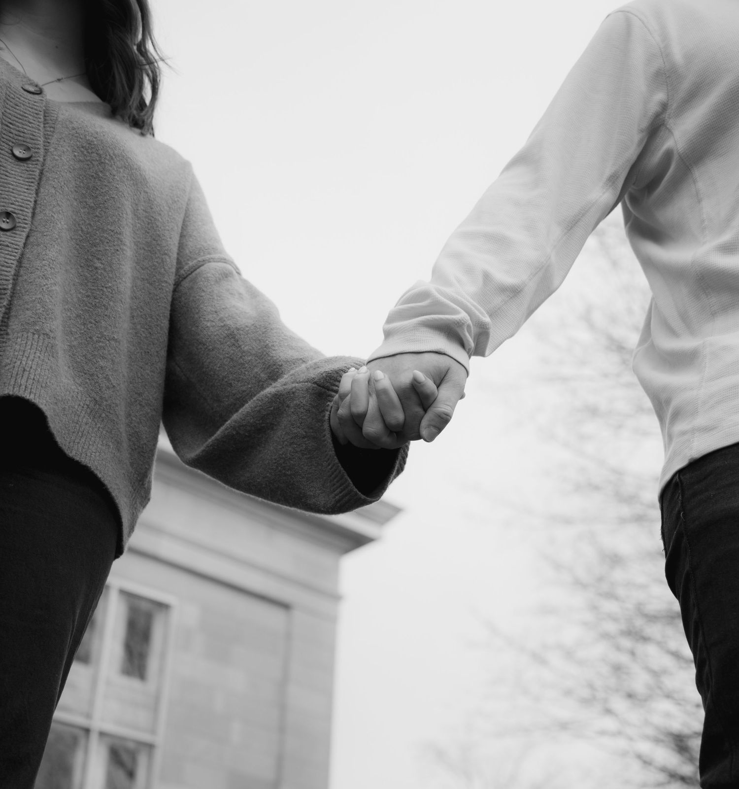 Black and white photo of a couple holding hands outdoors, viewed from behind, near a building.