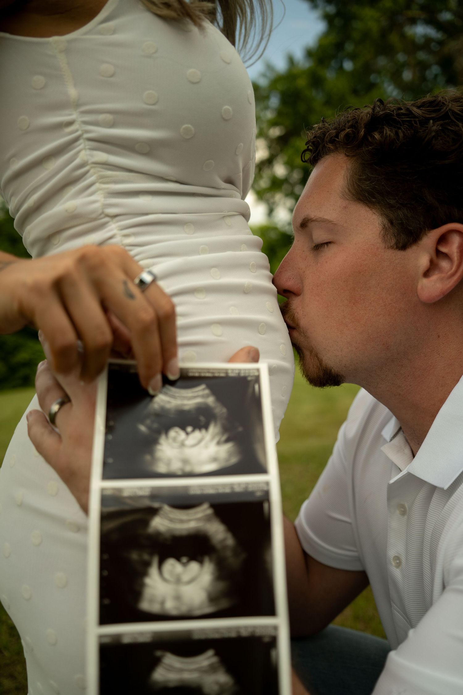 A man kisses a pregnant woman's belly outdoors while she holds ultrasound photos, both dressed in white.