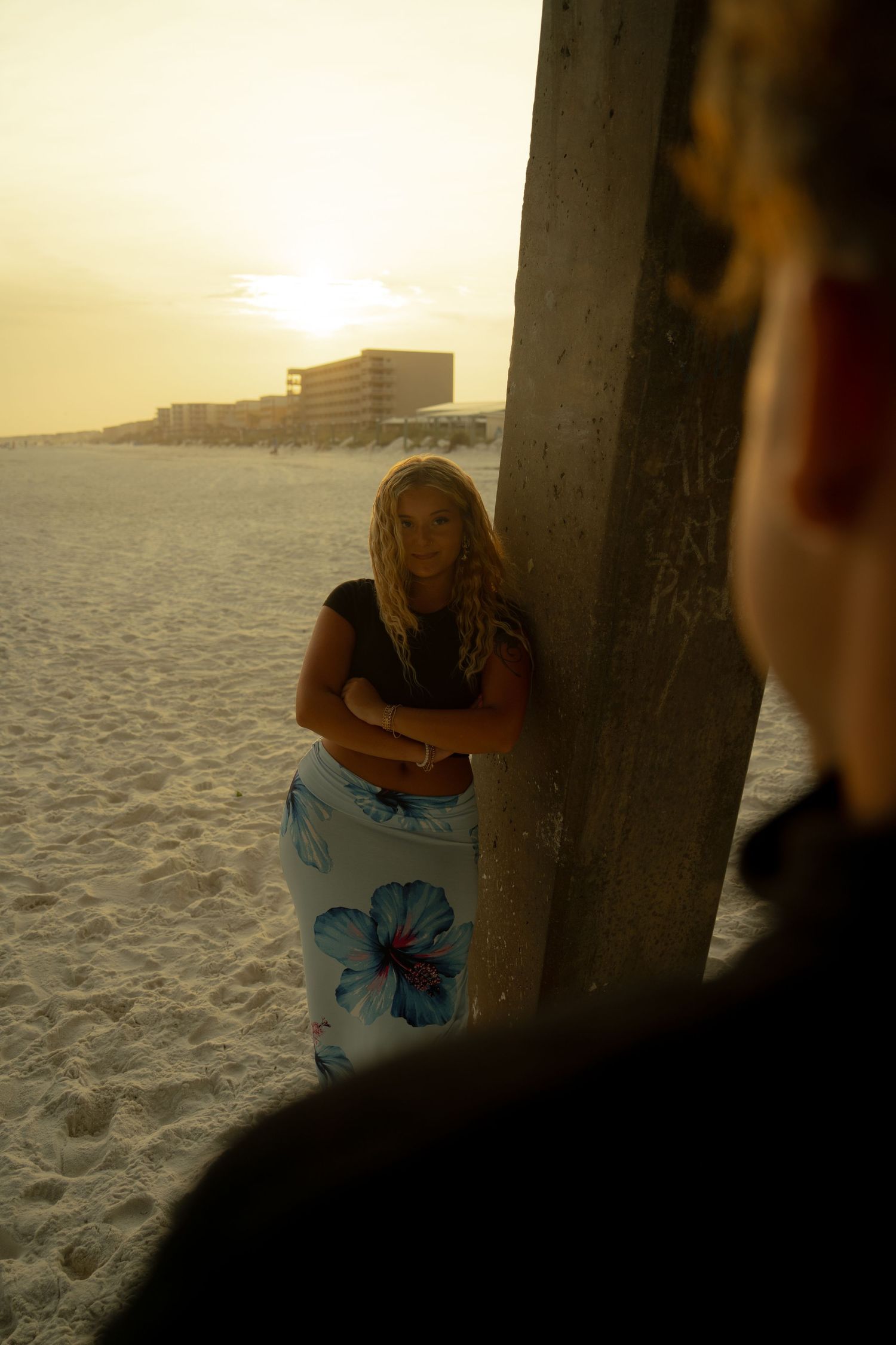 Blonde woman in floral sarong leans against pier pillar on white sand beach at golden sunset.