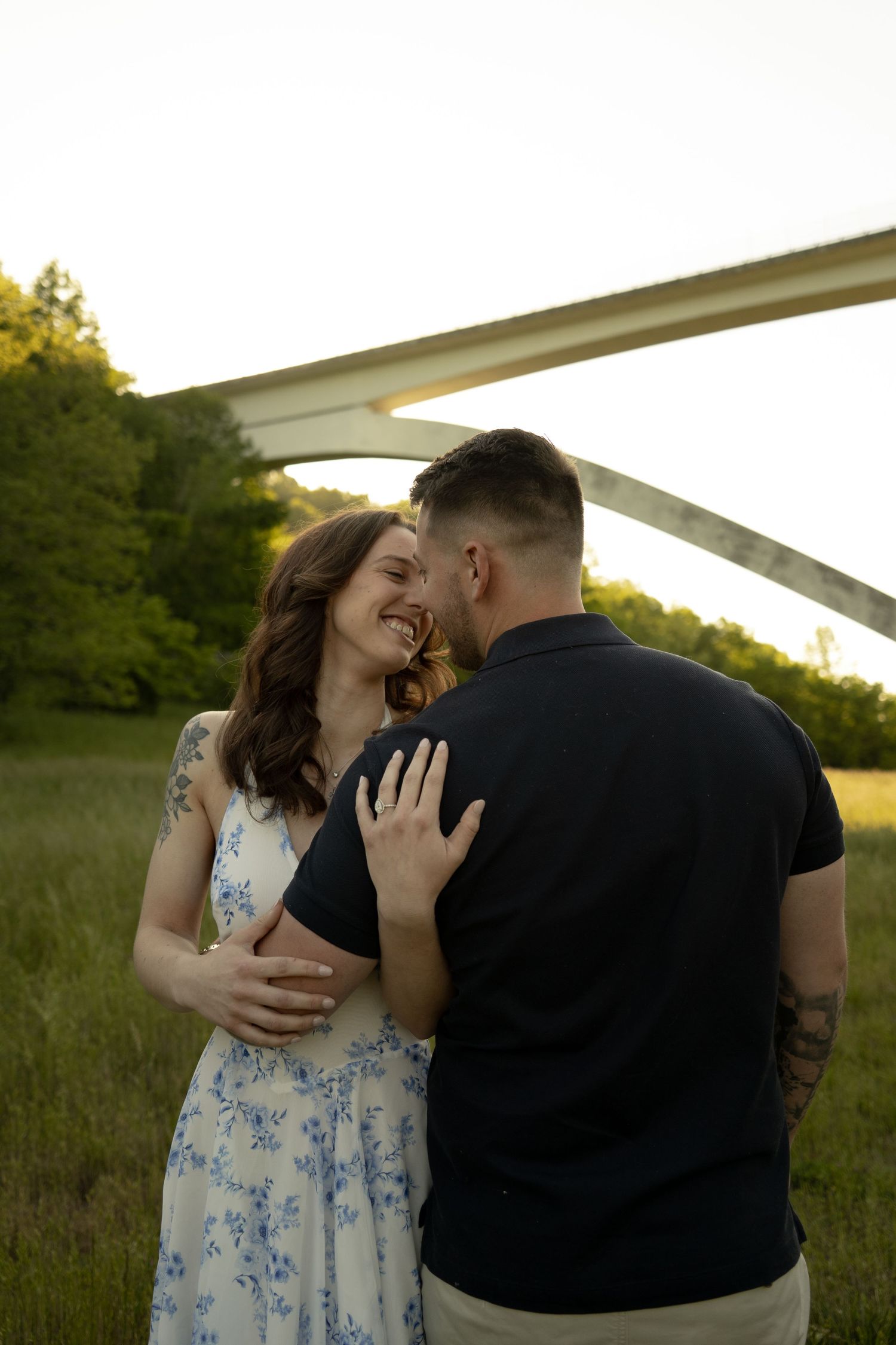 A smiling couple embraces in a green field at golden hour with an arched bridge and trees in the background.