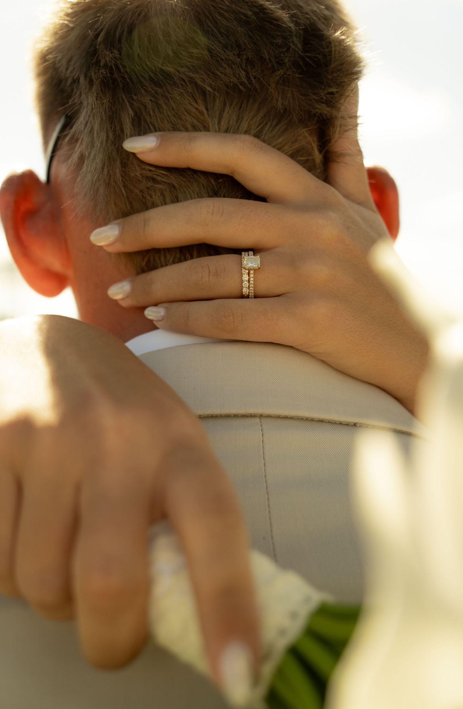 Close-up of a bride's hand with diamond engagement ring resting on groom's neck during wedding embrace.