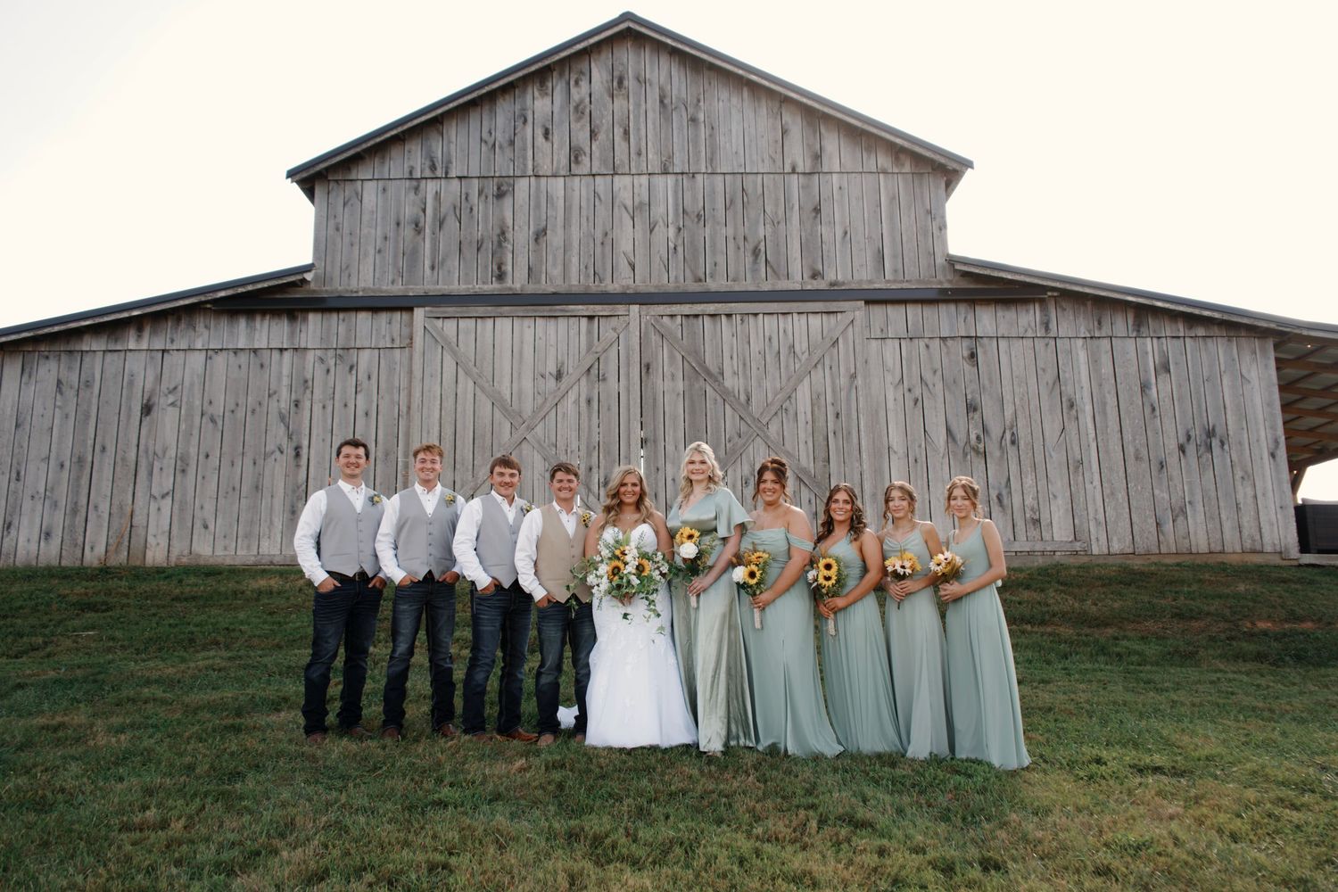 Wedding party poses in front of weathered gray barn for creative panoramic photo.