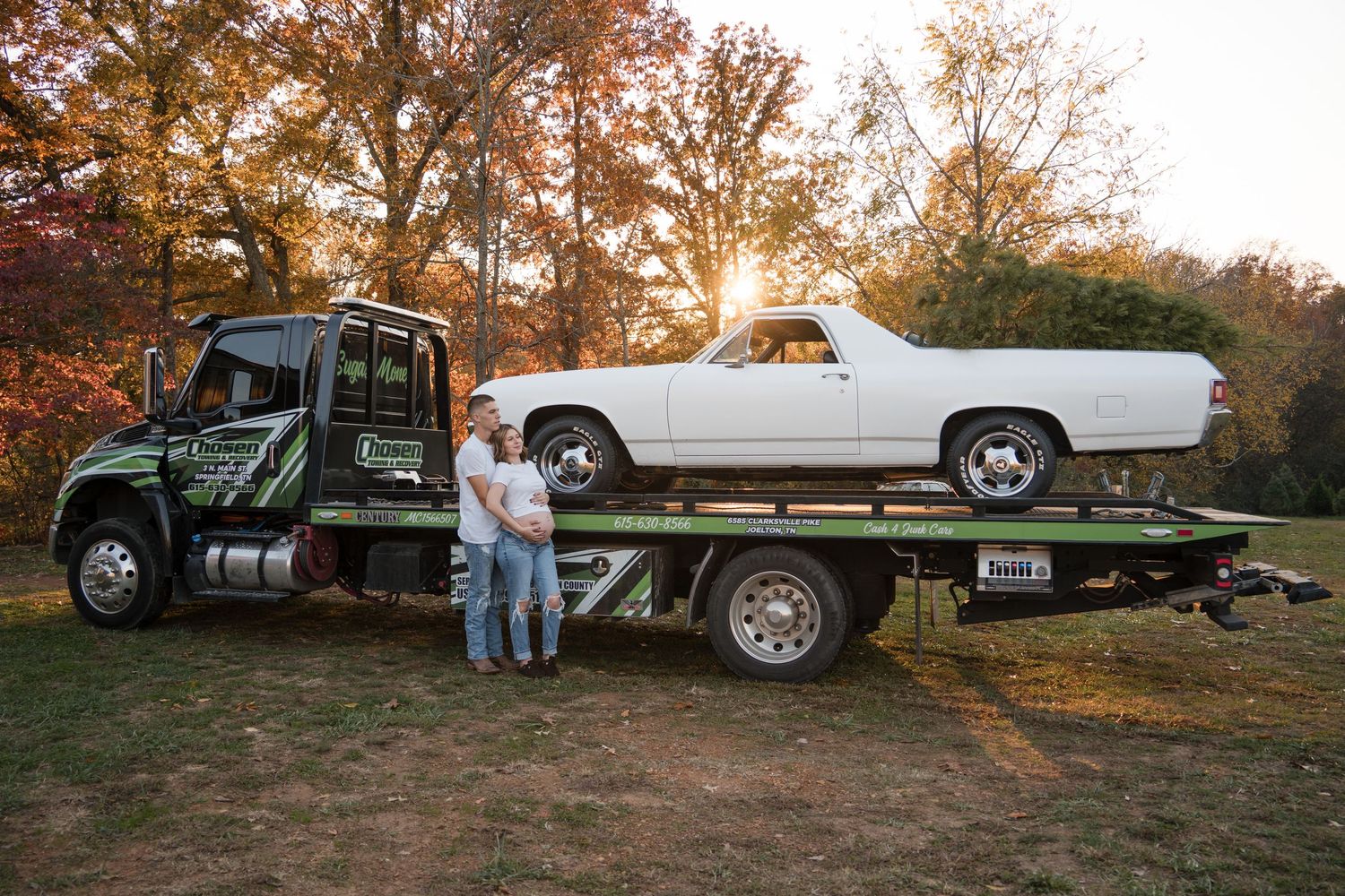 Vintage vehicles including a white classic car and flatbed truck are displayed on an autumn hill at dusk.