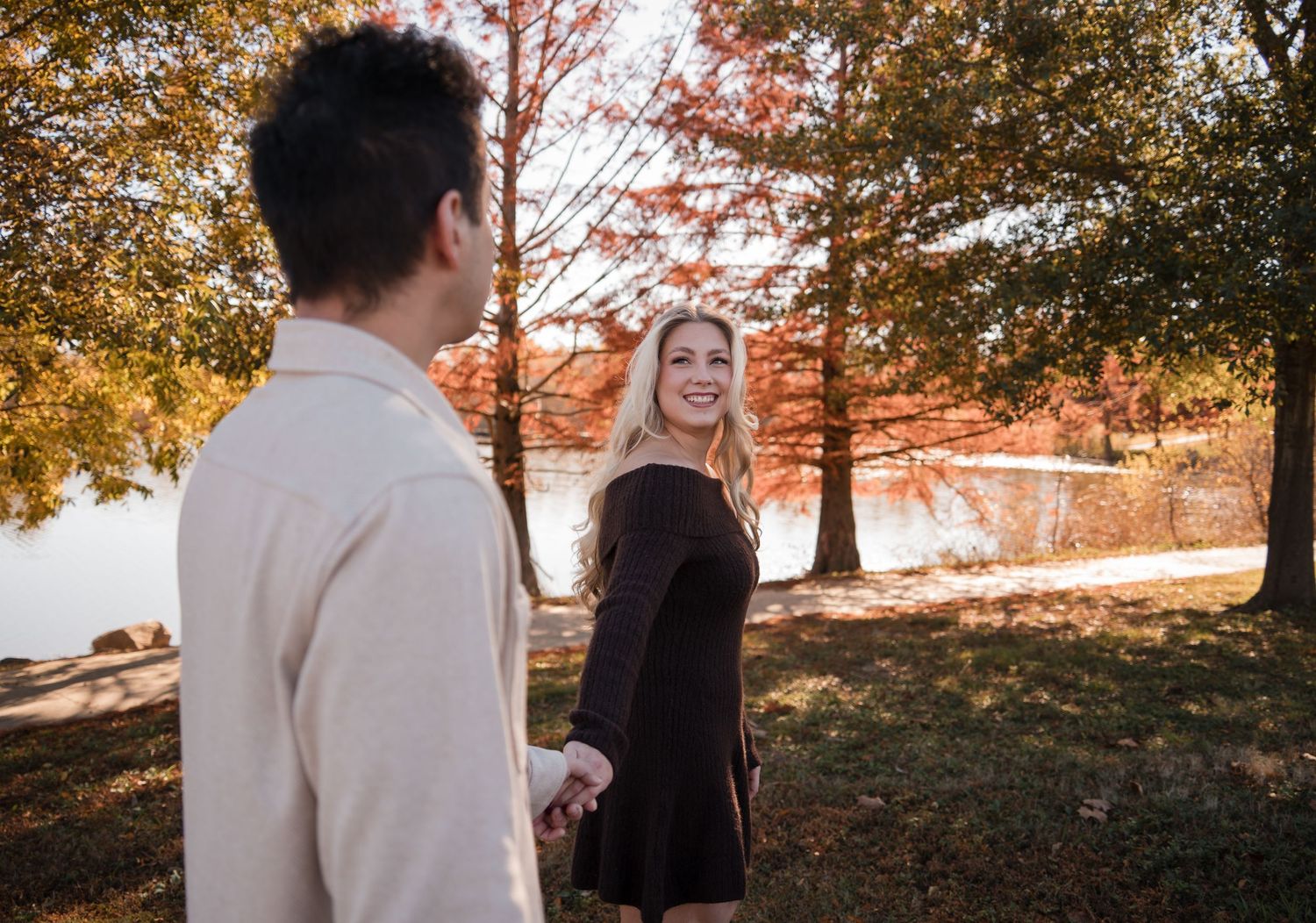 A couple holding hands in an autumn park during golden hour with fall foliage in the background.