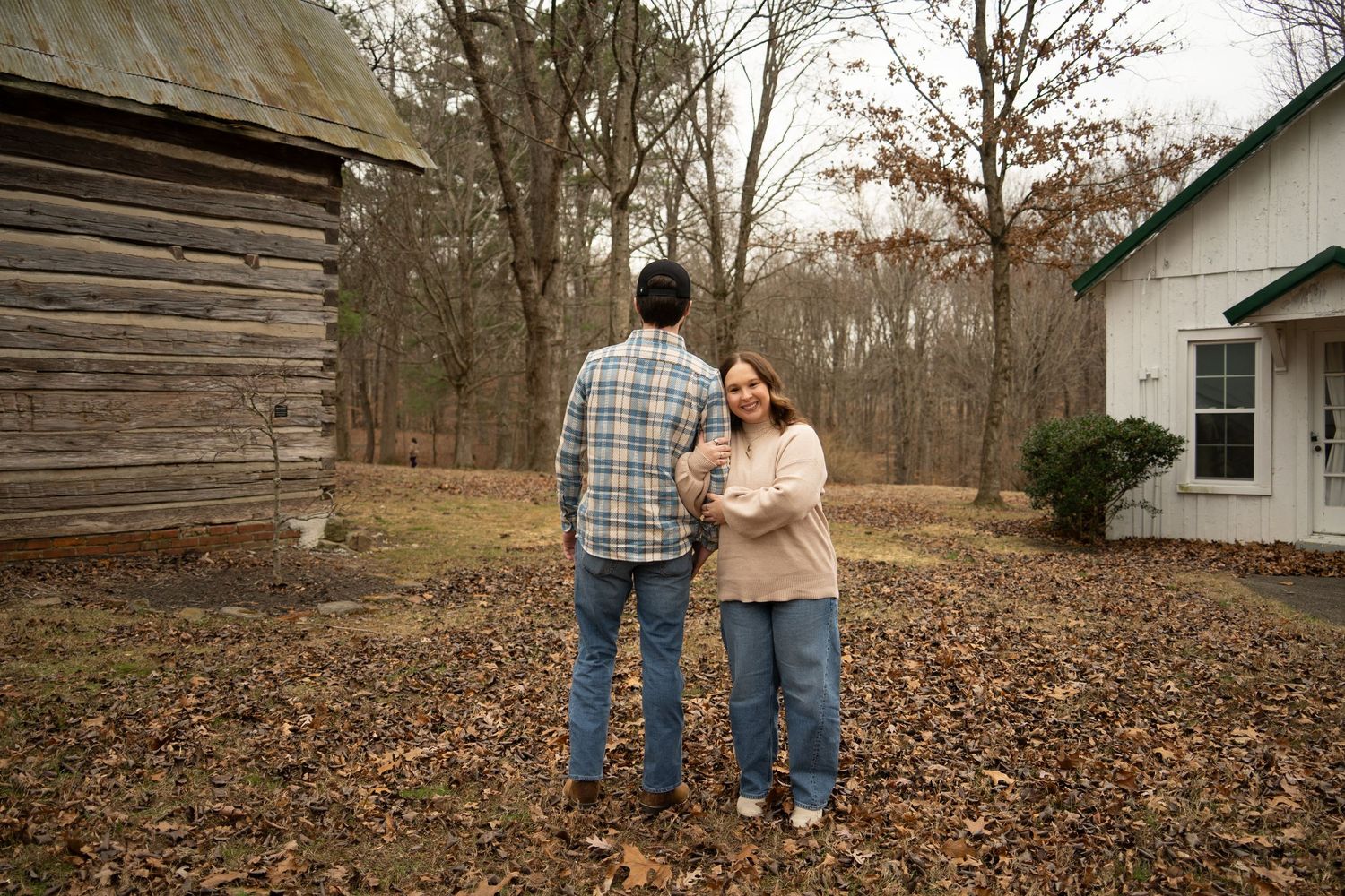 Couple stands together in autumn leaves outside rustic log cabin on a cloudy fall day.