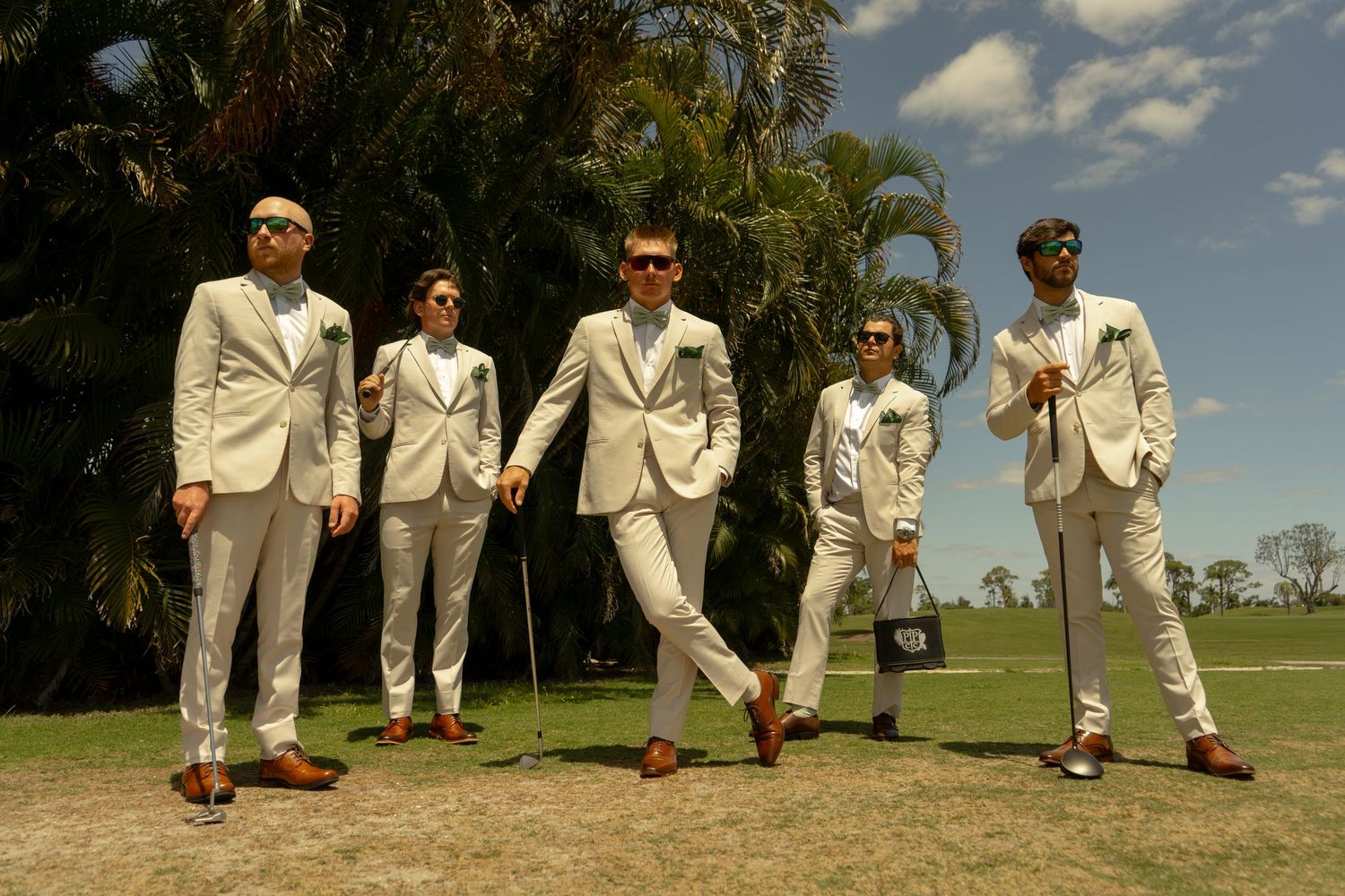Five men in matching white suits with brown shoes pose confidently on a golf course under a bright sunny sky.