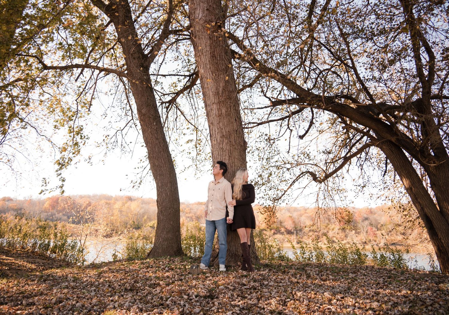 A couple stands beneath autumn trees on a hillside overlooking a river during golden hour.