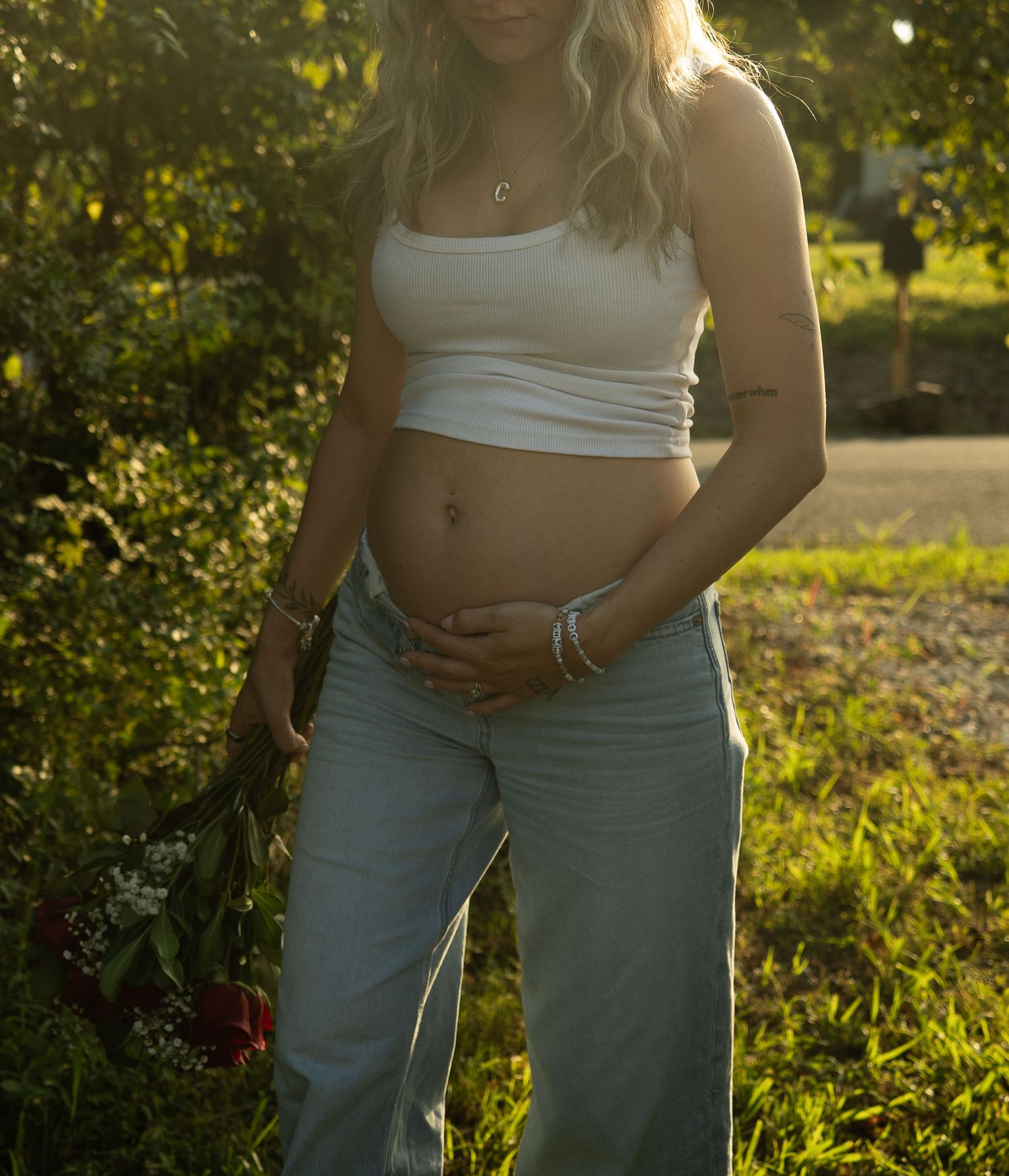 Pregnant woman in white crop top and jeans holding red roses outdoors in golden sunlight.