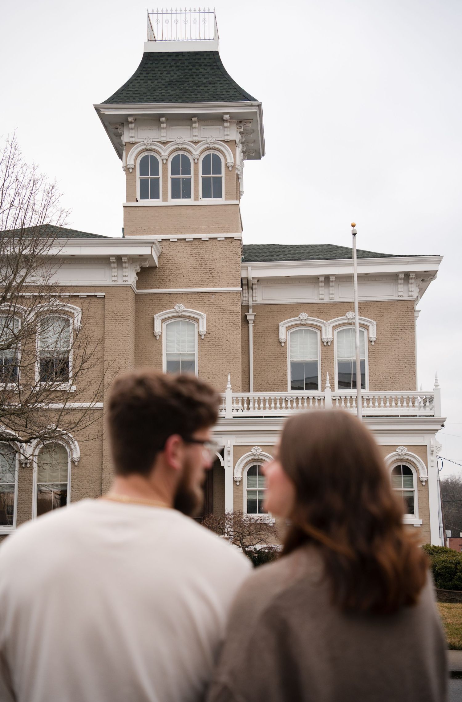Couple gazing at a historic Victorian brick building with a tower and ornate white trim on a cloudy day.