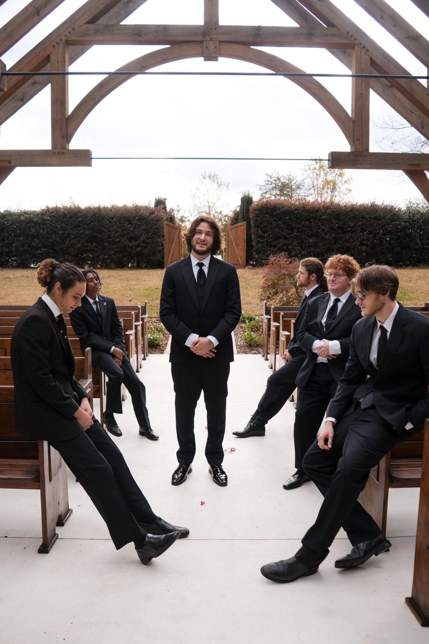 Groomsmen in black suits perform a playful kicking line dance sequence under a covered outdoor venue.