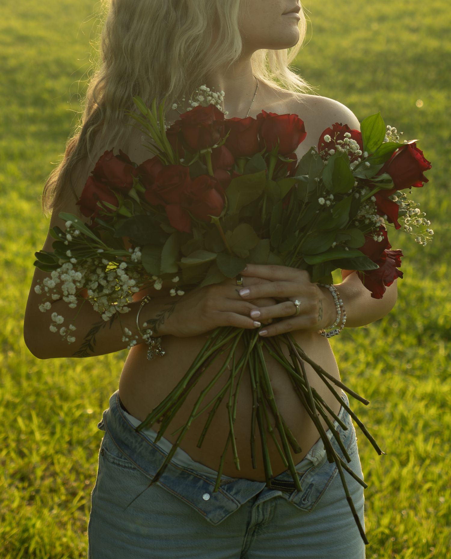 Woman holding large bouquet of red roses and baby's breath outdoors in green field, wearing jeans.